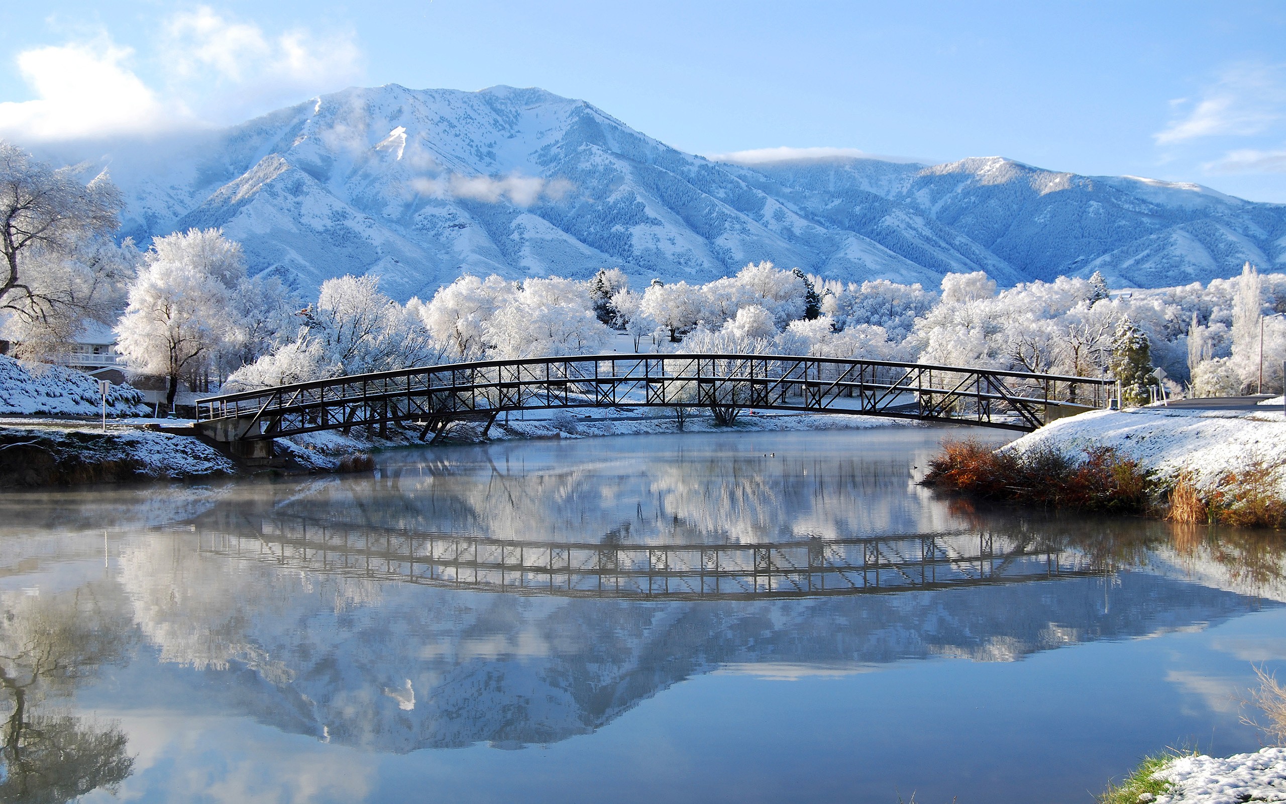 A serene winter landscape with a snow-covered bridge reflecting in a calm lake, set against a backdrop of majestic, snow-capped mountains and frost-covered trees