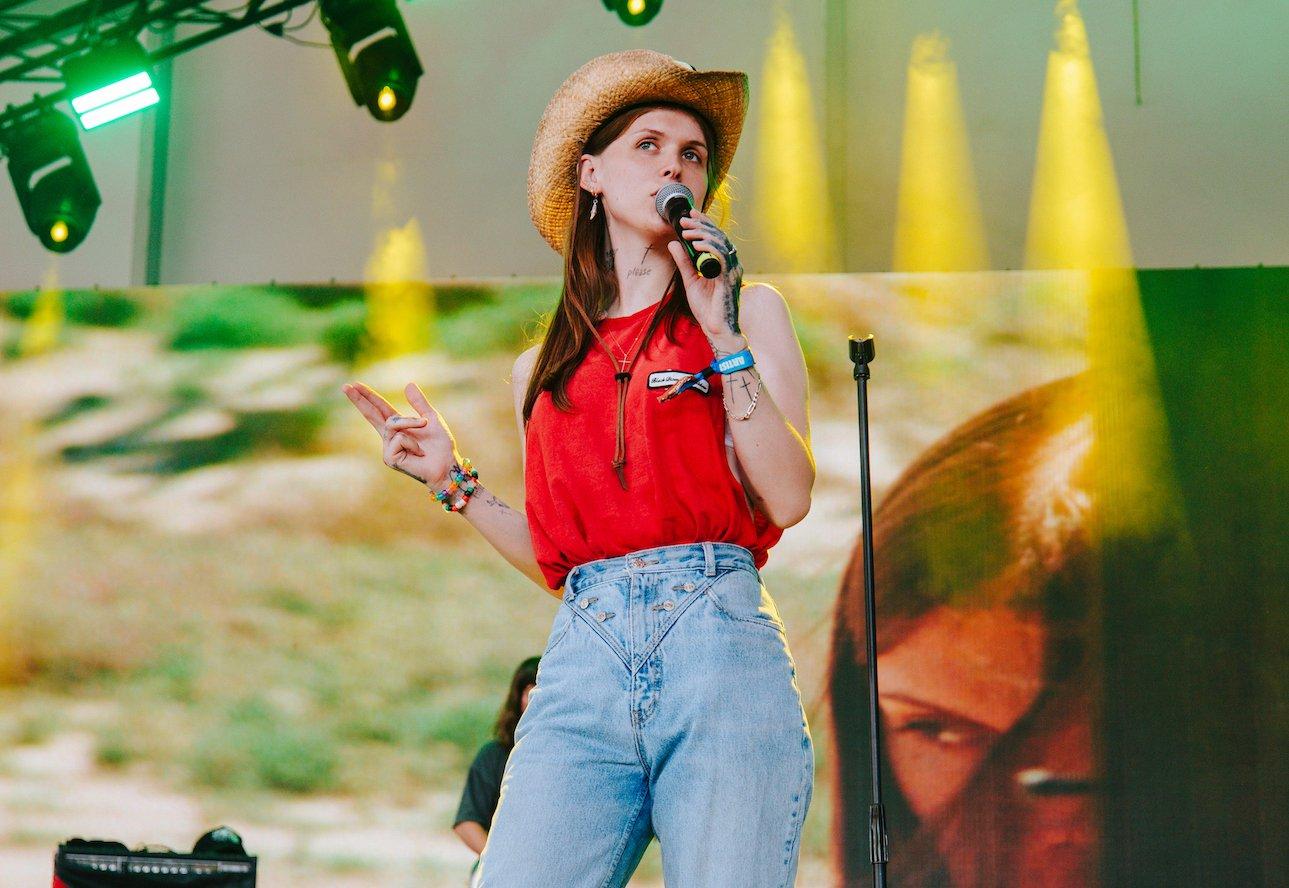 A singer performs on stage, wearing a straw cowboy hat, red sleeveless top, and light denim jeans. They hold a microphone in one hand and gesture with the other