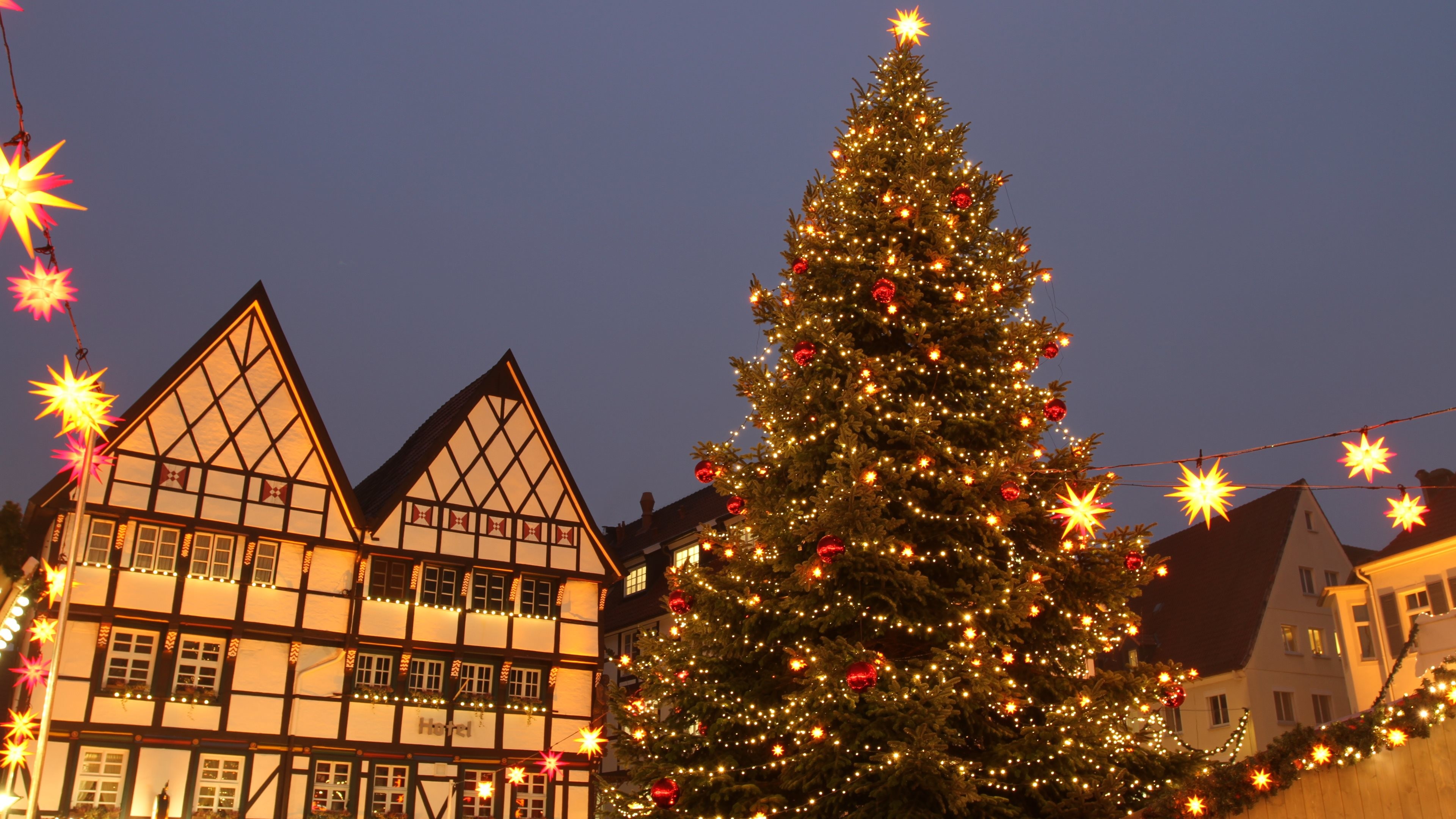 A towering Christmas tree, adorned with twinkling lights and ornaments, stands majestically in a town square, creating a magical holiday atmosphere against the backdrop of traditional half-timbered houses