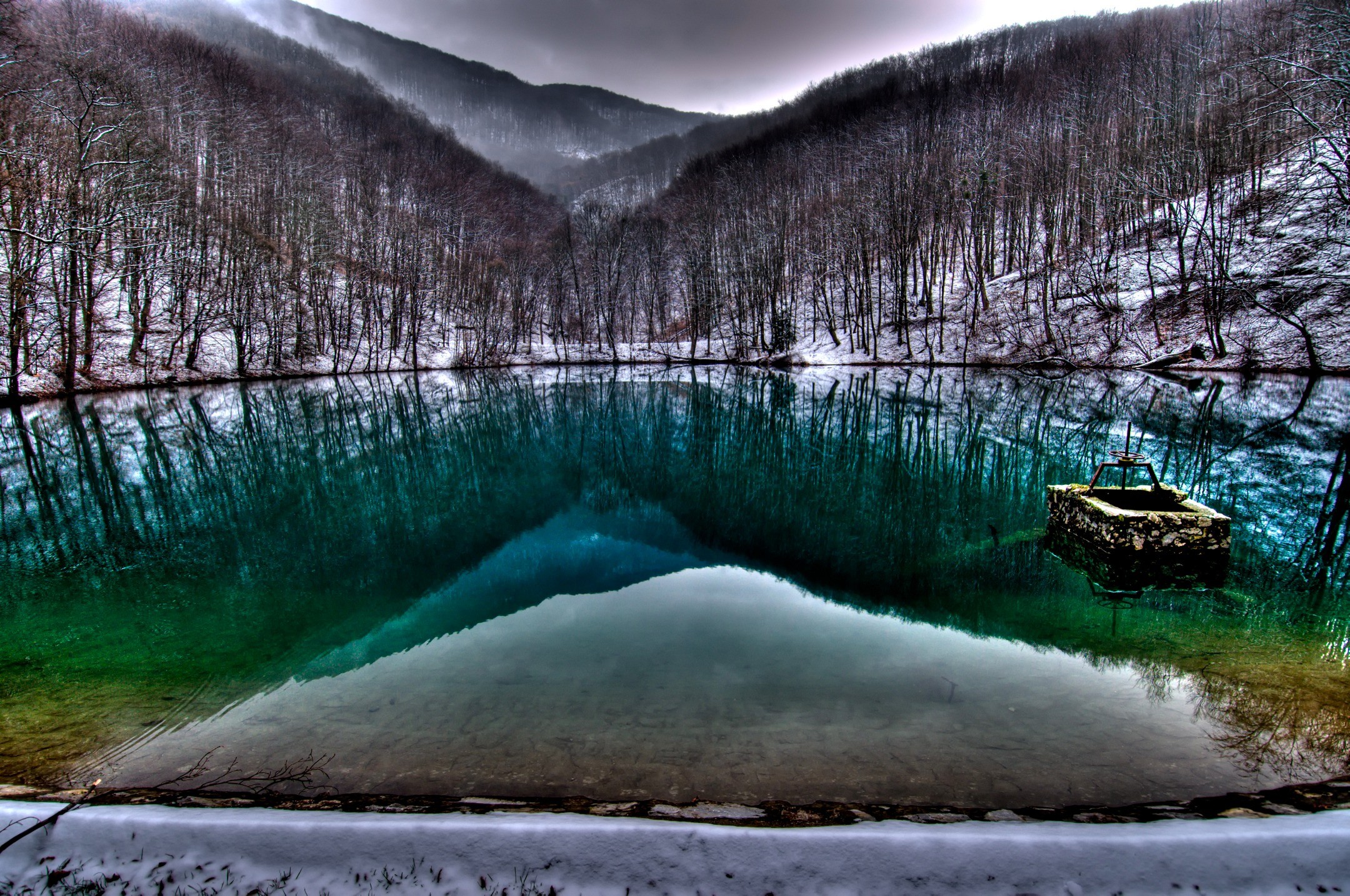 A serene winter landscape with a crystal-clear lake reflecting the snow-dusted trees and mountains