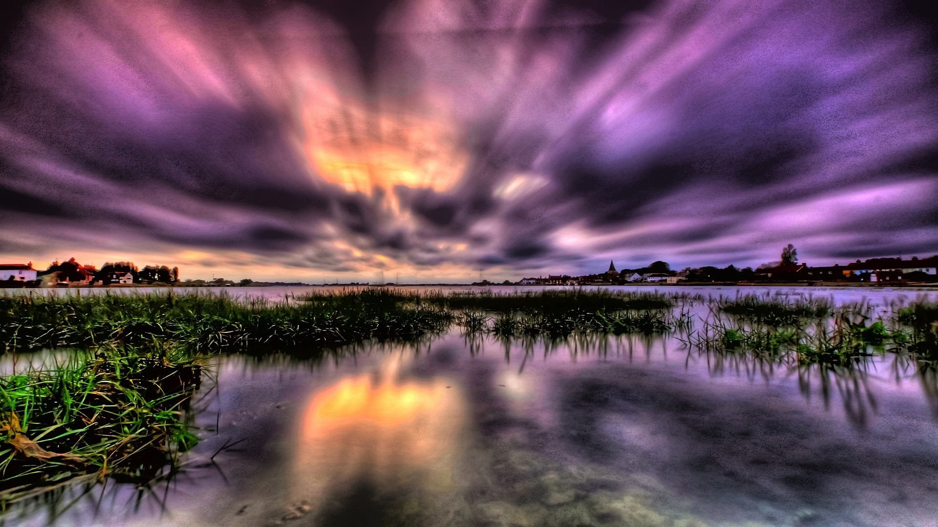 A breathtaking sunset over a serene lake, with vibrant purple and orange hues reflecting in the calm water. Patches of grass emerge from the shallows, adding to the tranquil beauty of the scene
