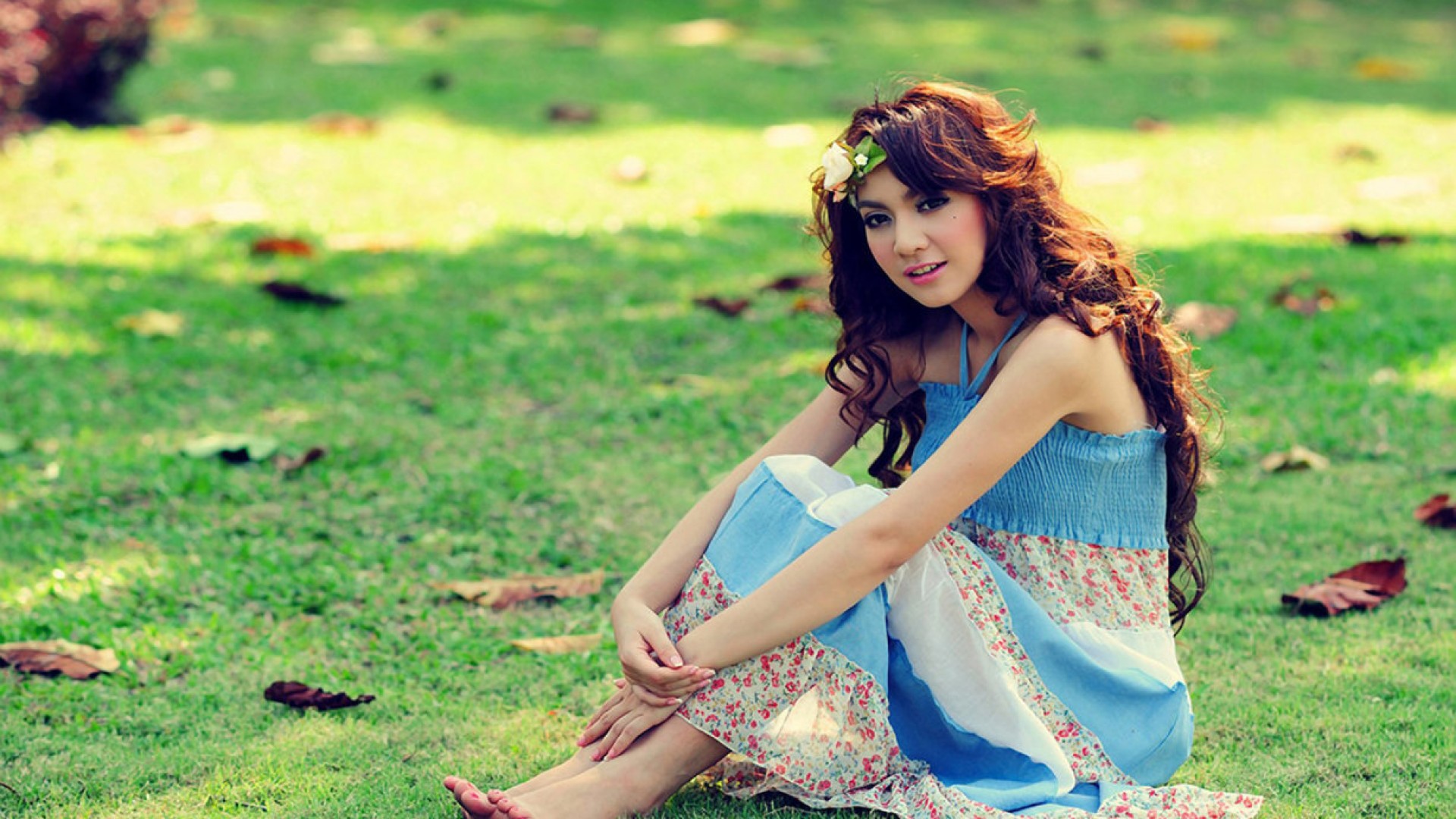 A young woman sits gracefully on a grassy lawn, wearing a floral sundress and a flower in her hair
