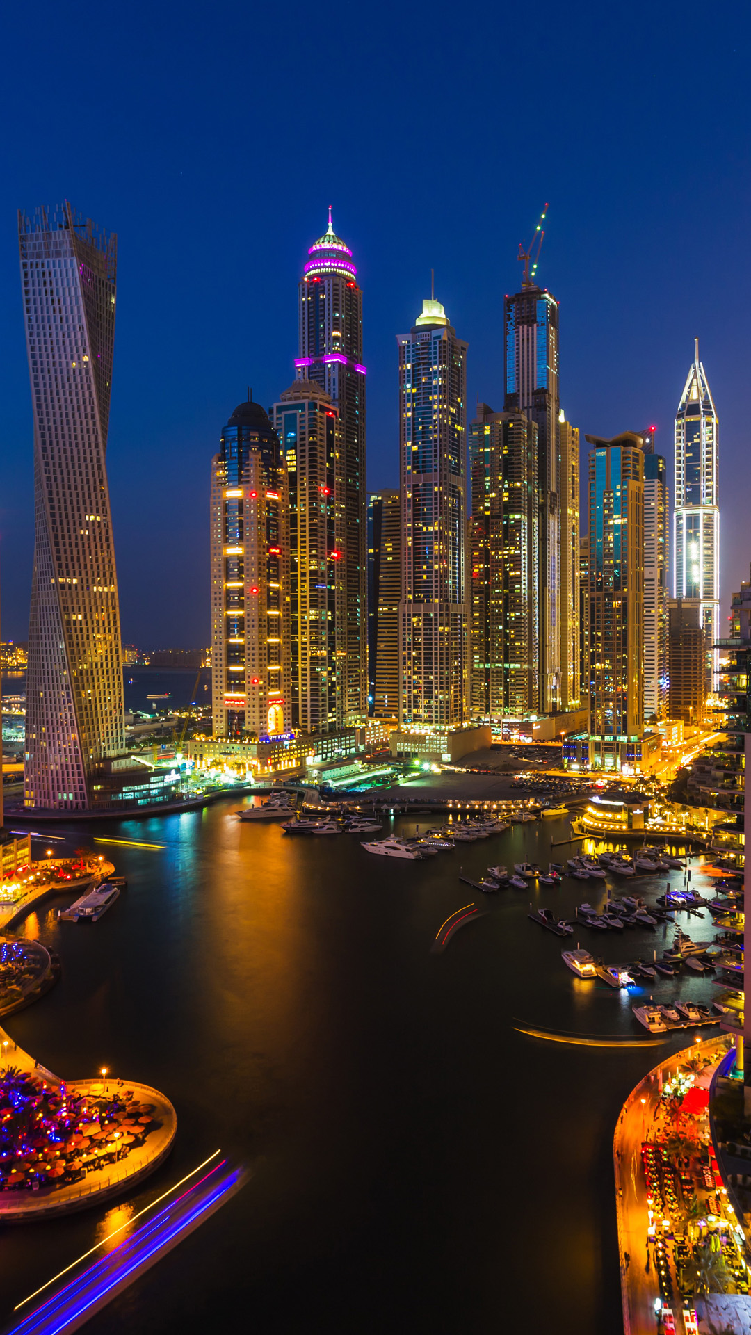 Dubai Marina cityscape at night, with illuminated skyscrapers and a harbor full of boats. The long-exposure photo captures light trails from moving boats and cars
