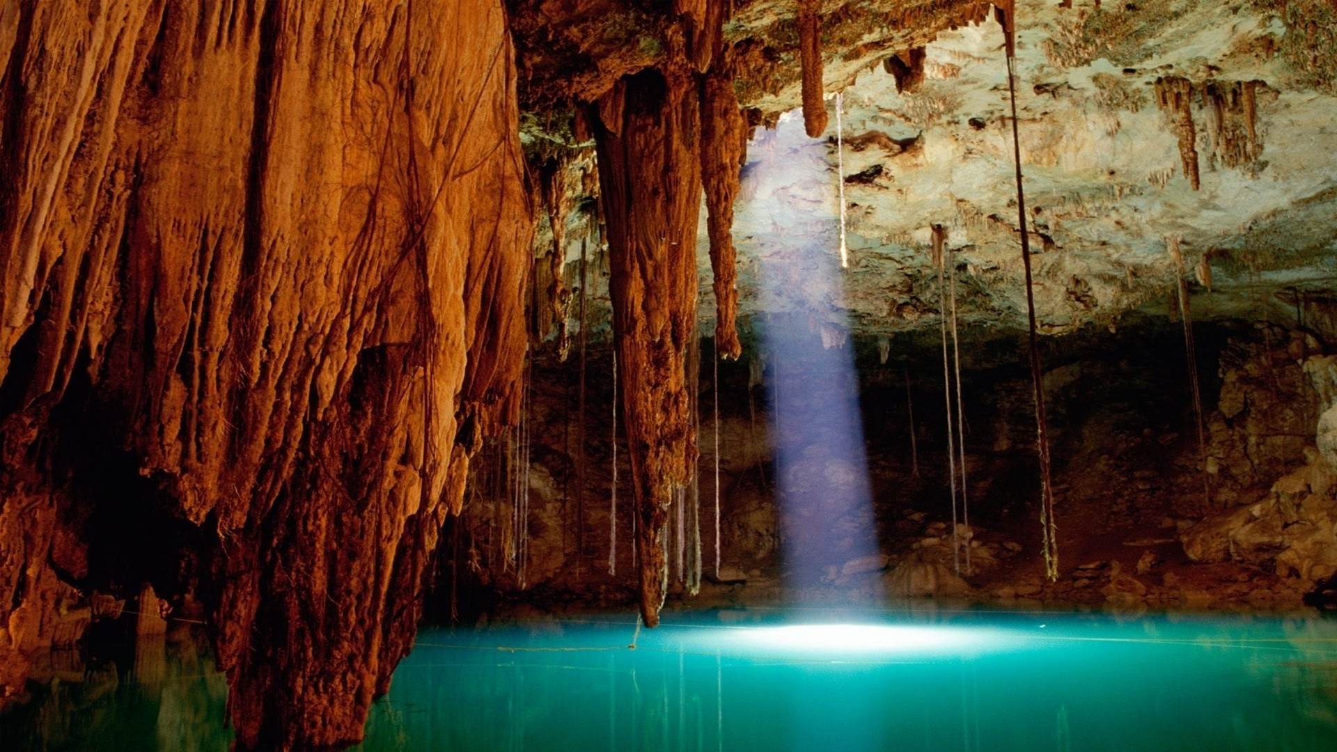 Sunlight illuminates a turquoise underground cenote in a cave, stalactites hanging from the ceiling, creating a mystical atmosphere