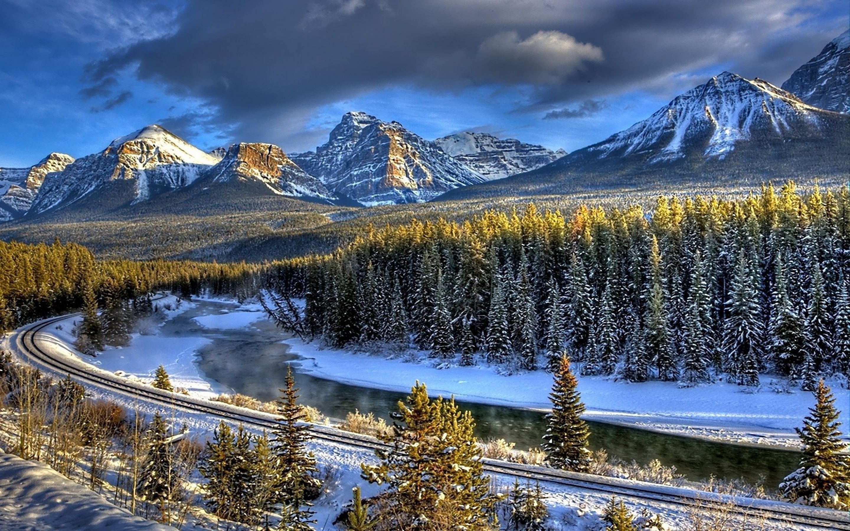Scenic winter landscape with snow-covered mountains, a frozen river, and a railroad track winding through a pine forest
