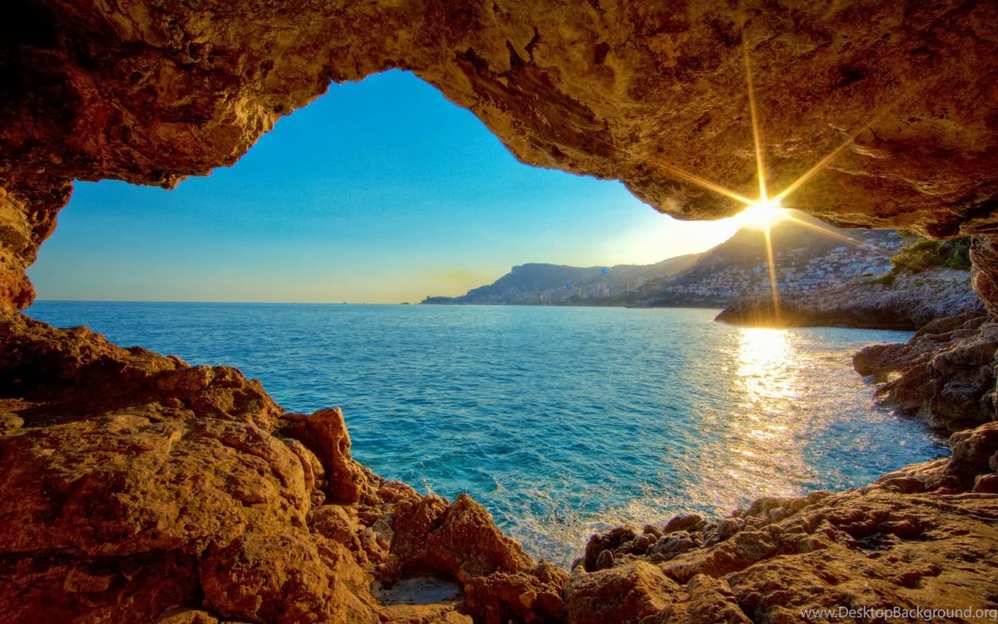 Golden sunlight streams into a sea cave, illuminating the turquoise waters and a coastal town in the distance