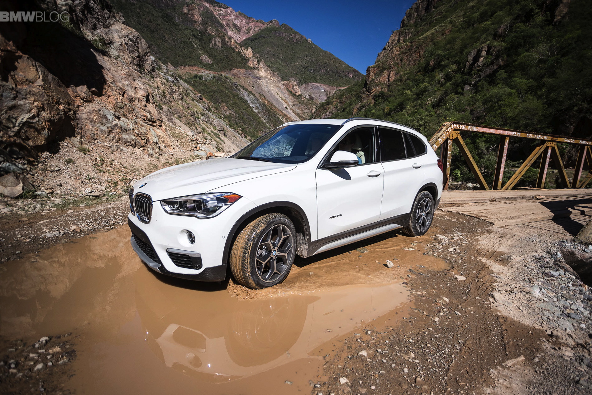 White BMW X1 SUV parked in a muddy puddle on a dirt road, with a rusty bridge and rocky mountains in the background