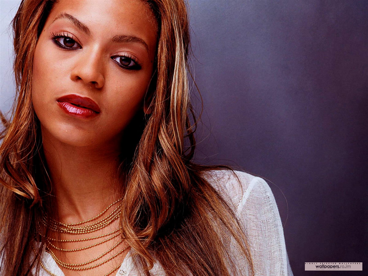 Close-up of Beyoncé, a famous American singer, songwriter, and actress, with her long, wavy brown hair cascading over her shoulders. She is wearing a white top and a delicate gold necklace, her expressive eyes and subtle makeup adding to her captivating presence