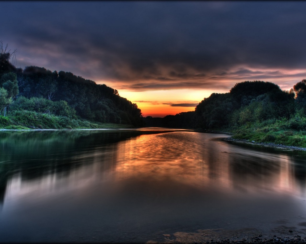 Serene sunset over a calm river reflecting the colorful sky and bordered by lush green trees