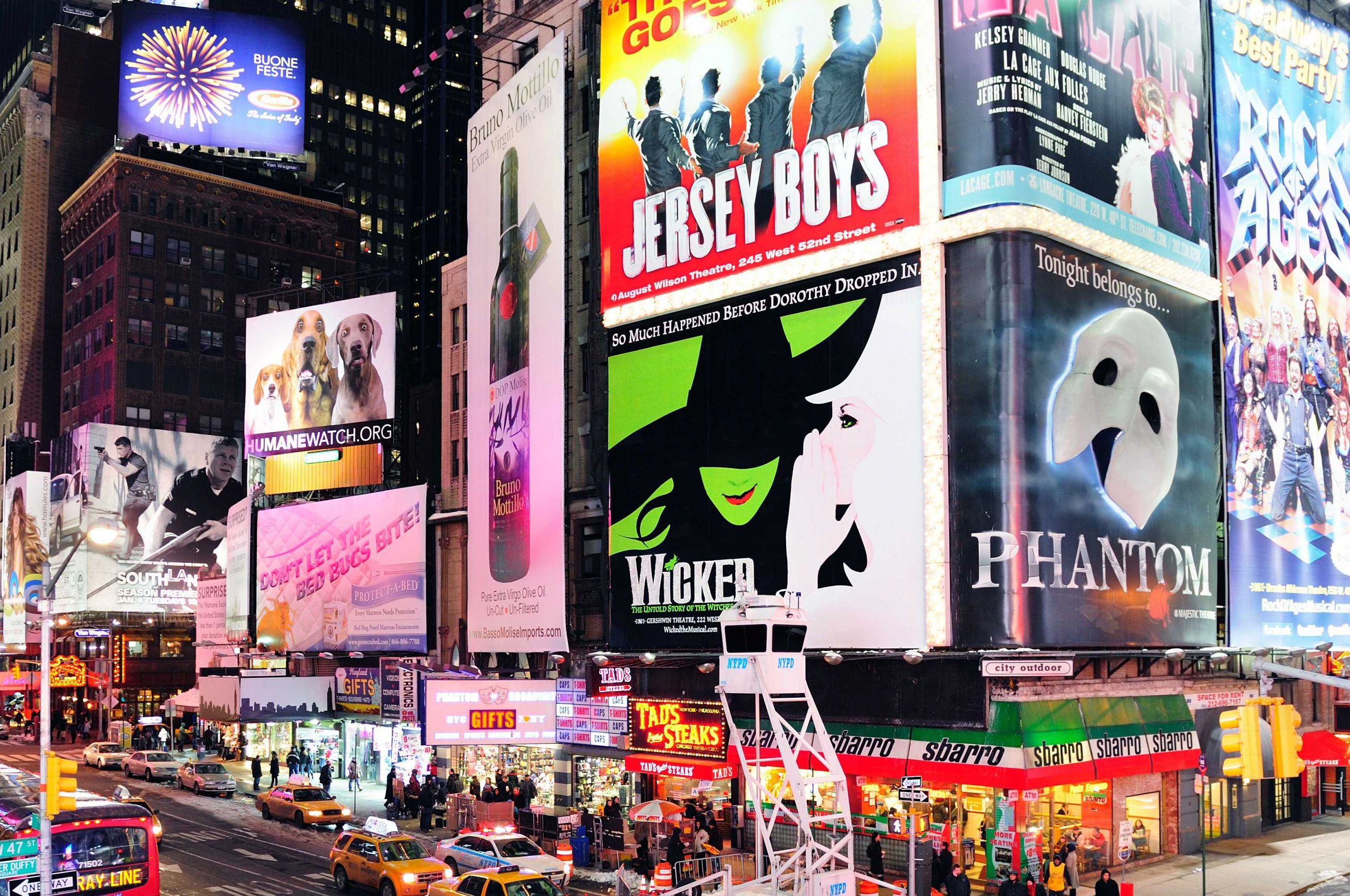 A vibrant night scene in Times Square, New York City, showcasing illuminated billboards advertising Broadway shows like Jersey Boys, Wicked, and Phantom of the Opera, amidst bustling city life