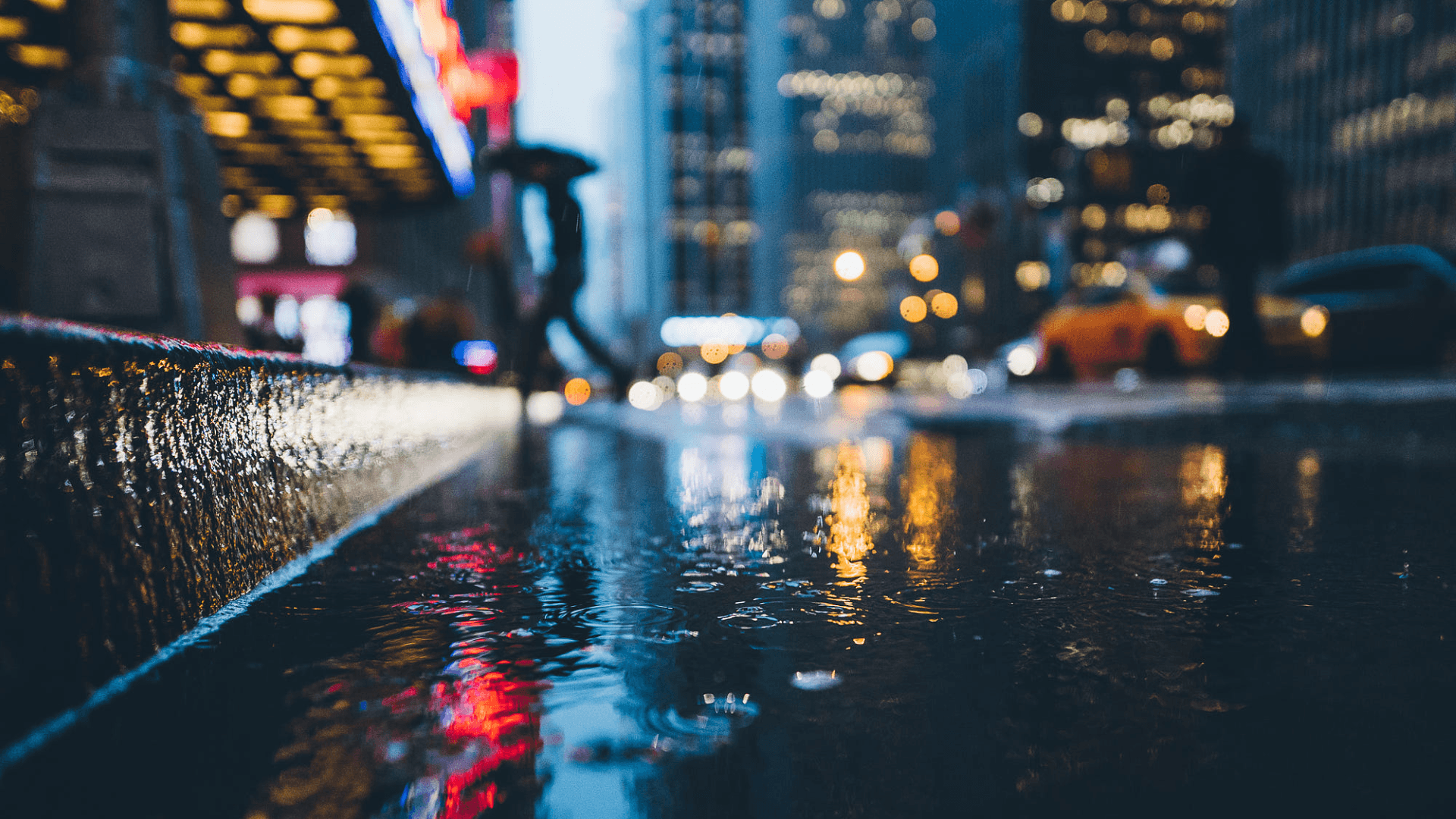 Close-up of a rain puddle on a city street at night, reflecting the blurred lights of cars and buildings