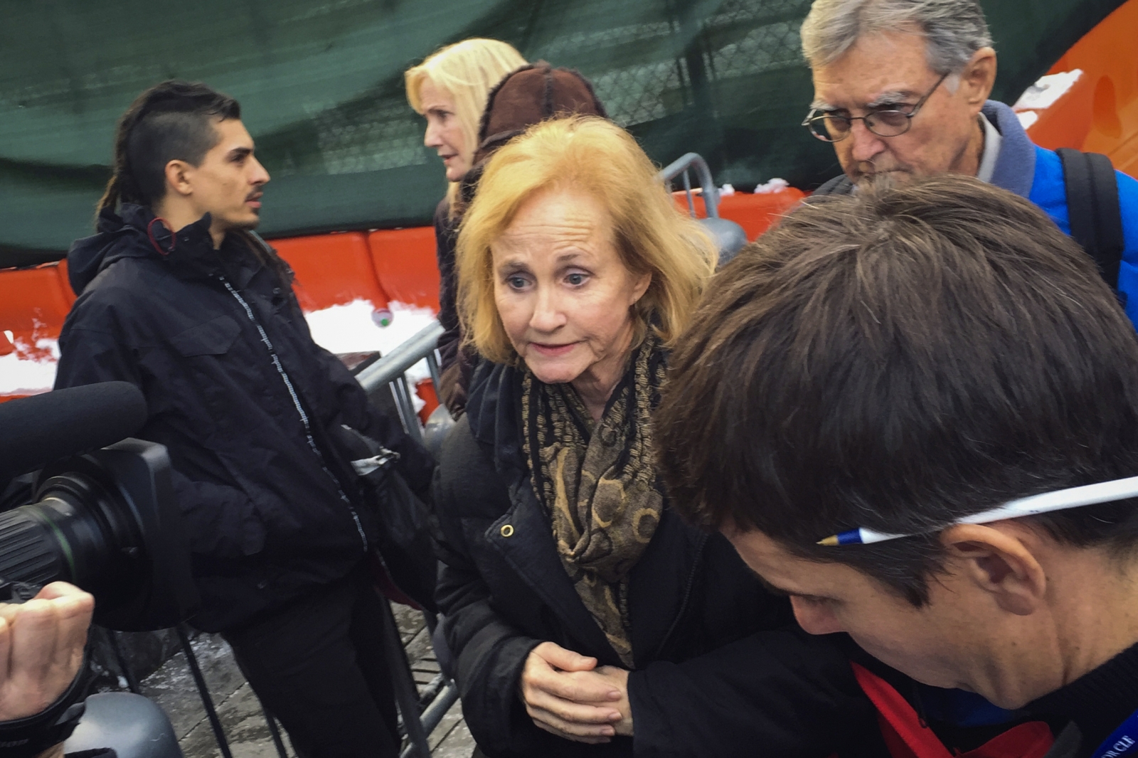 A group of people, possibly including journalists and members of the public, gathered near an outdoor venue. The focus is on a woman with light hair who appears to be giving a statement or being interviewed