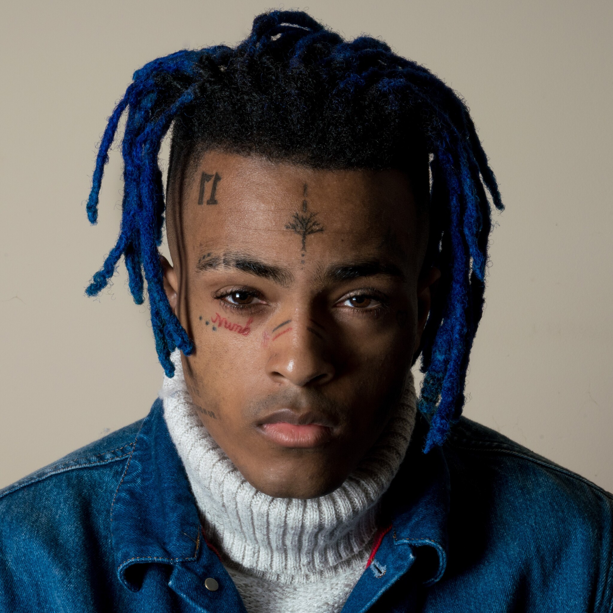 Close-up portrait of XXXTentacion with blue dreadlocks, face tattoos, and a denim jacket