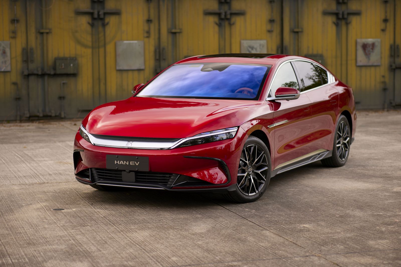 A red BYD Han EV parked in front of a weathered industrial backdrop