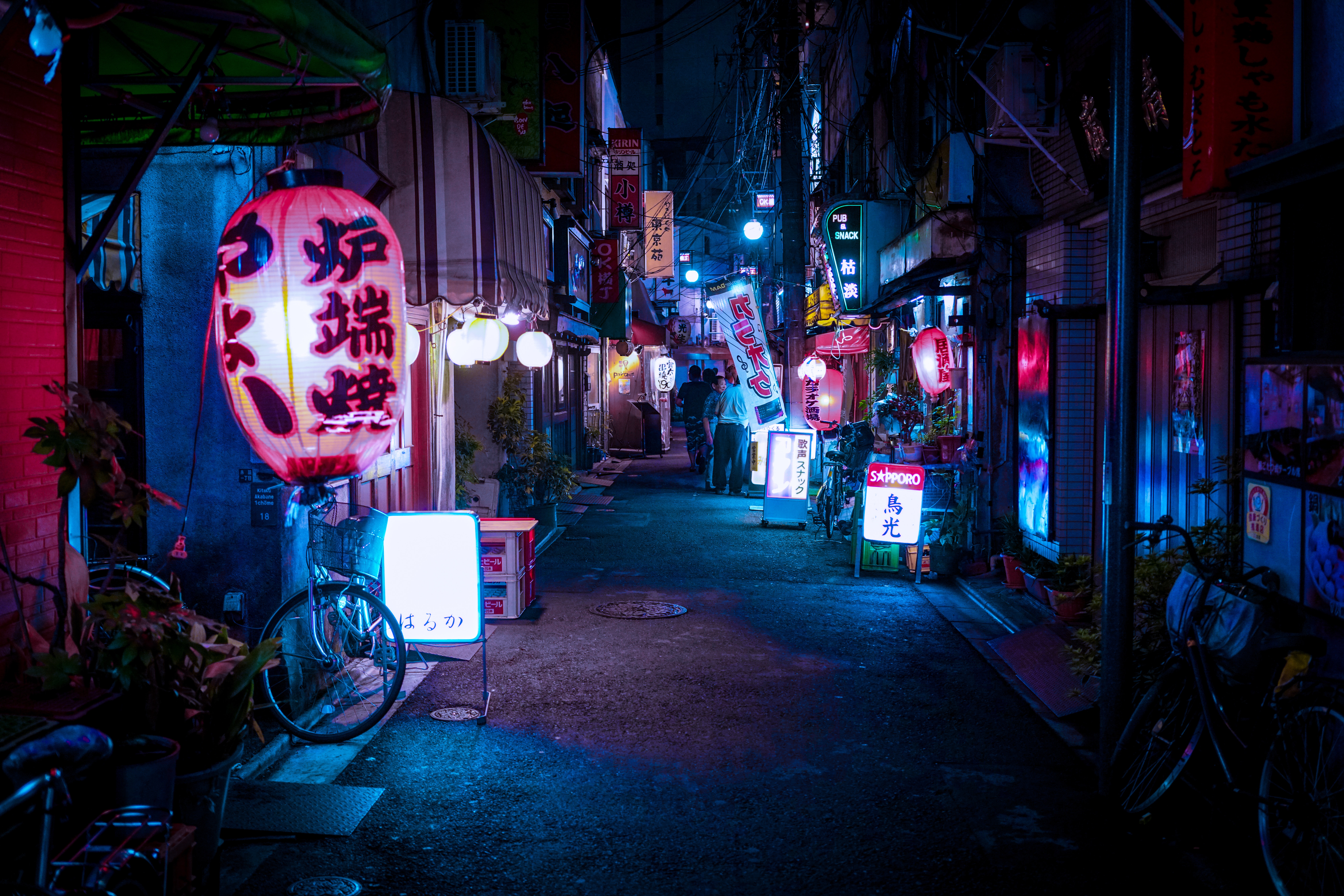 A vibrant night view of a narrow Japanese alleyway, illuminated by glowing lanterns, neon signs, and various shop lights. People stroll through the lively scene