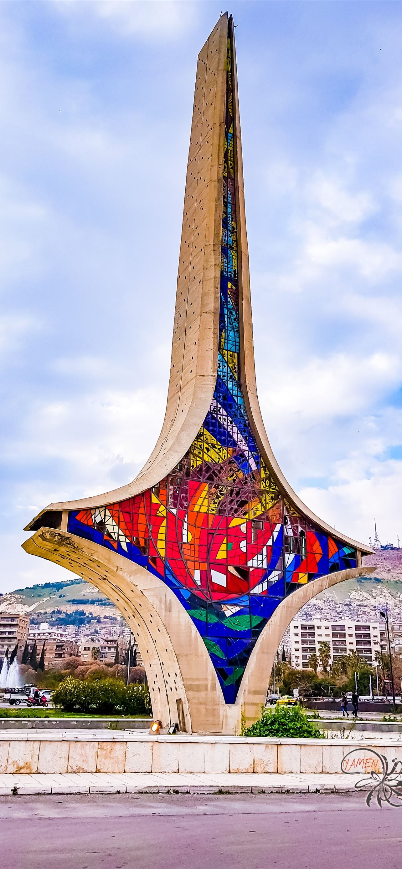 A vibrant stained-glass monument stands tall against a cloudy sky in Syria, showcasing a unique blend of art and architecture