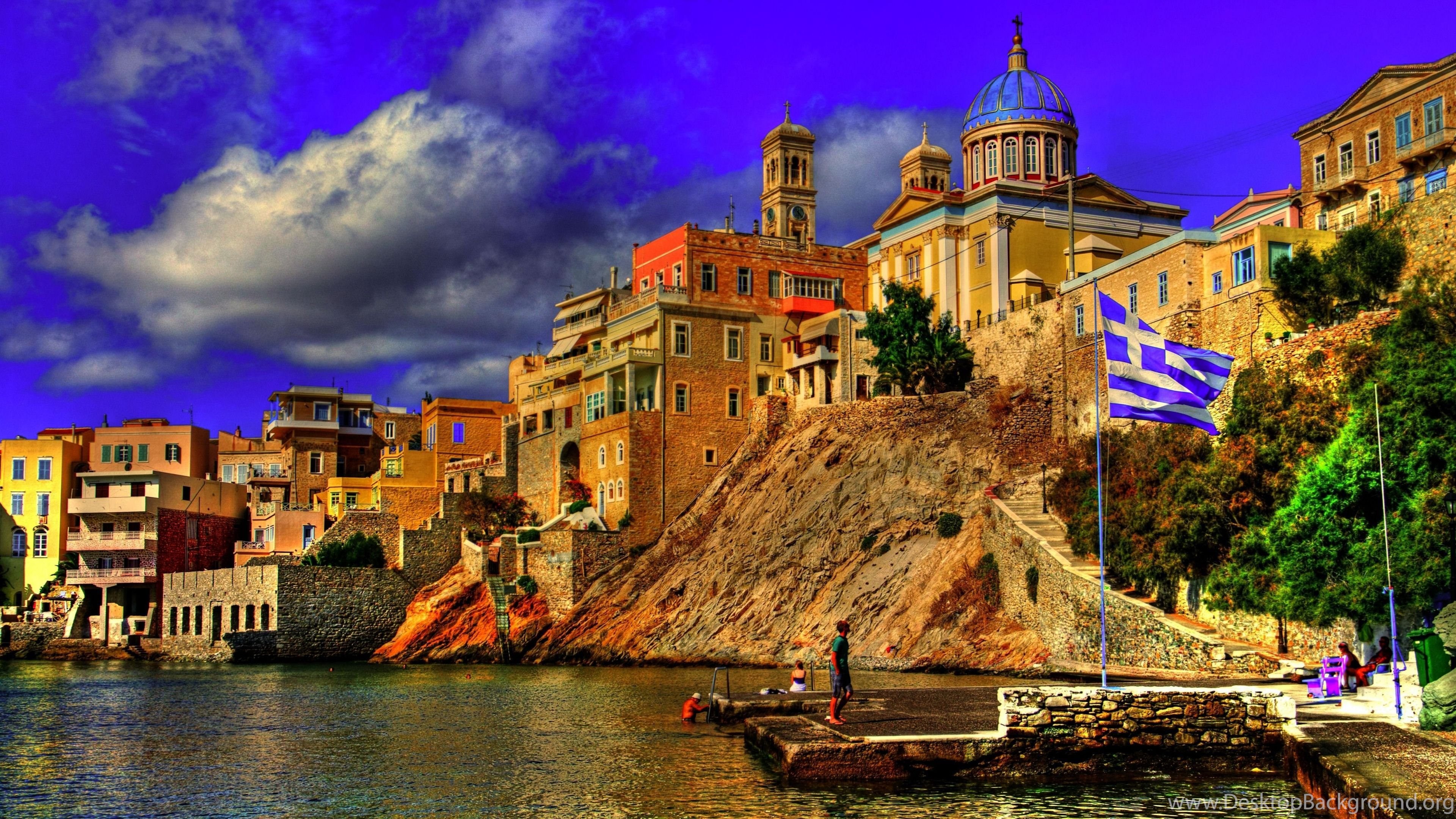 Scenic view of a Greek coastal town with colorful buildings, a domed church, and the Greek flag waving proudly
