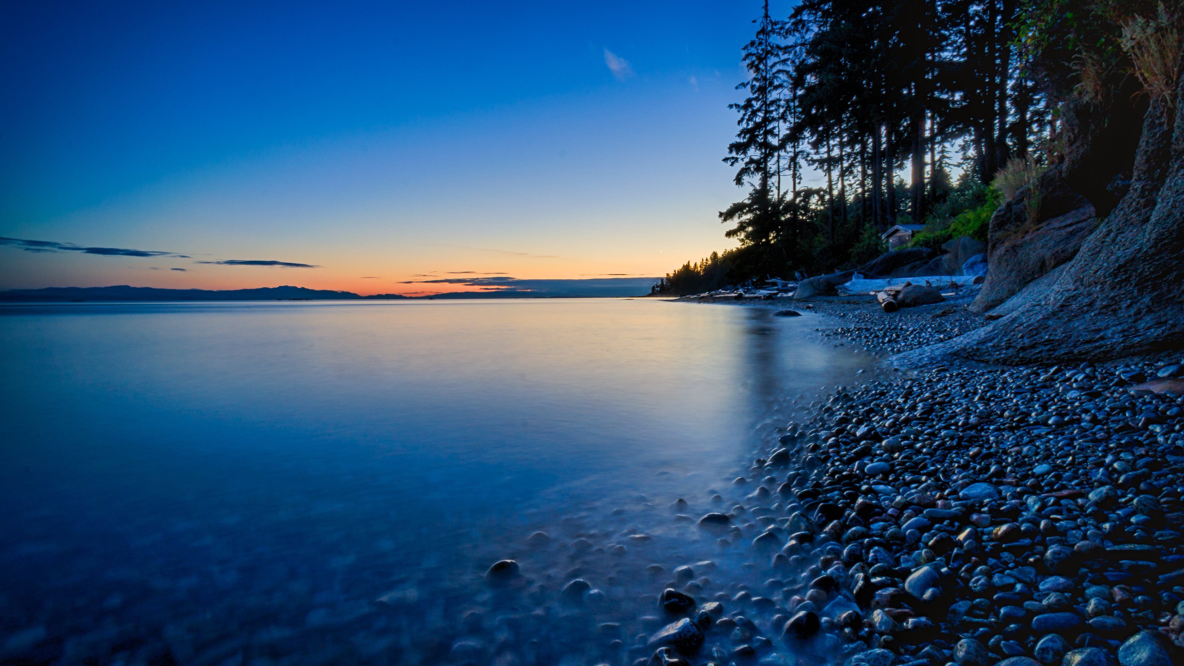 A breathtaking 4K wallpaper capturing the serene twilight over a tranquil pebble beach. The smooth, dark-blue water reflects the vibrant hues of the setting sun, while a line of silhouetted trees adds a touch of mystery to the scene