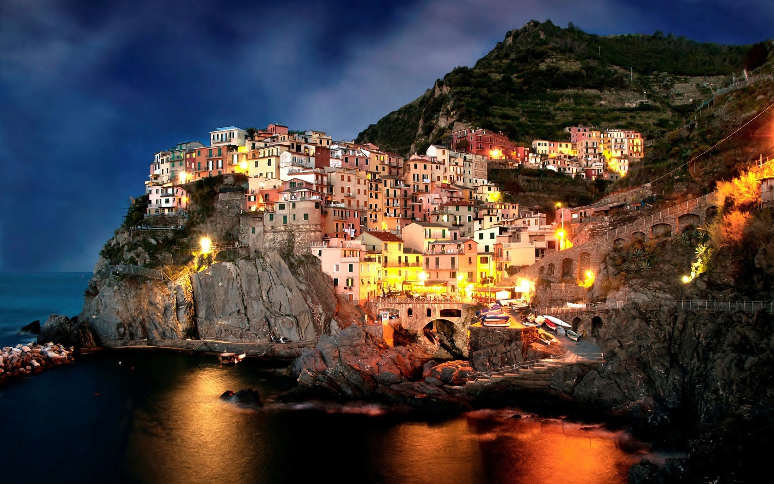 A breathtaking 4K night view of Manarola, Italy, a picturesque village nestled on cliffs overlooking the Ligurian Sea. The colorful houses glow warmly against the dark rocks and blue sky, creating a magical atmosphere