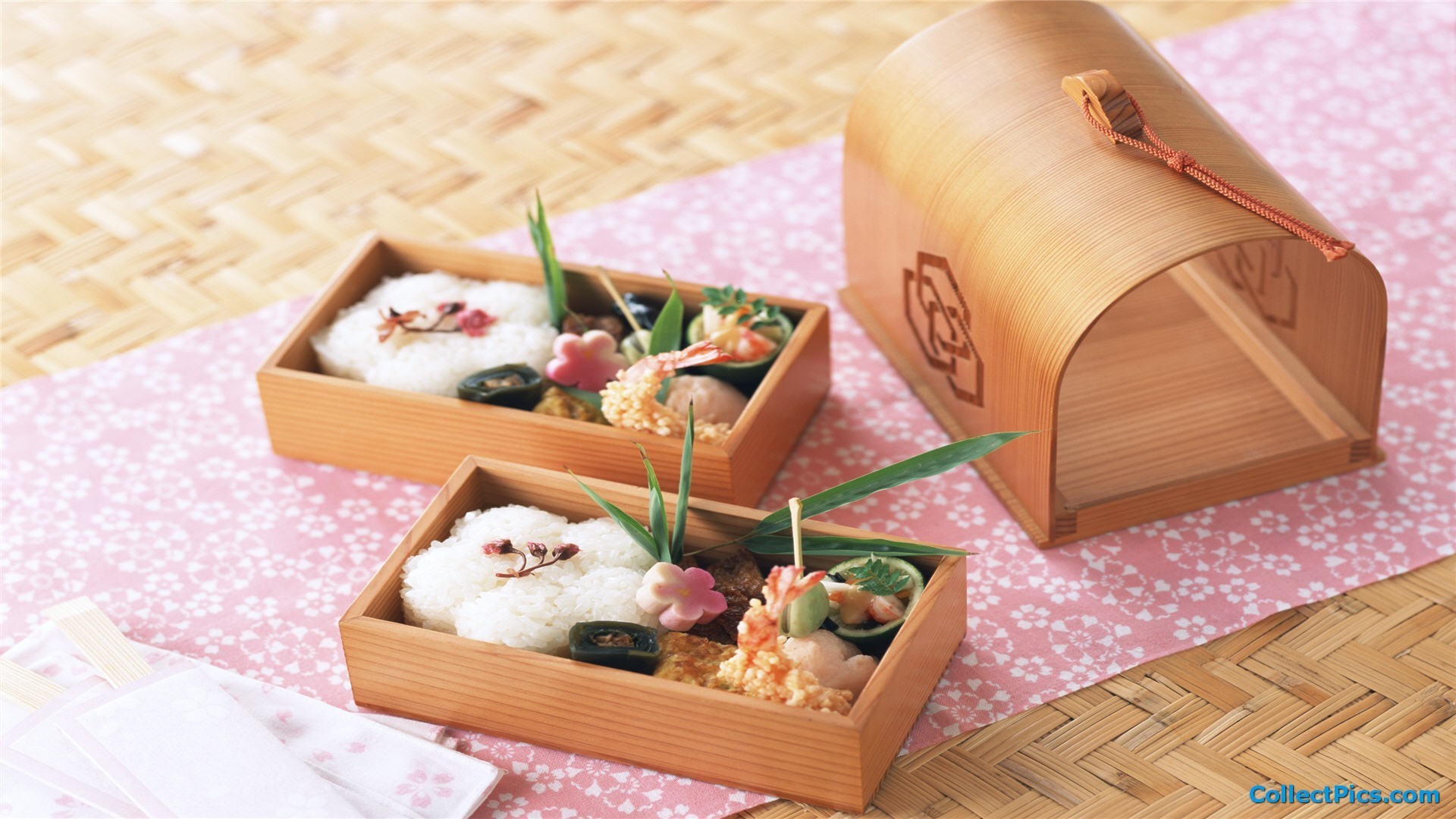 Two wooden bento boxes filled with decorative Japanese food, placed on a pink floral cloth over a woven bamboo mat. A larger, curved wooden container sits behind them