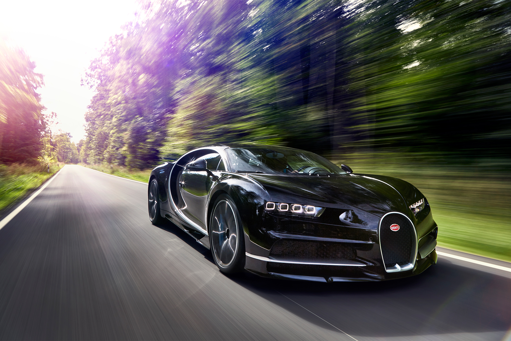 A black Bugatti Chiron Super Sport speeds down a paved road lined with blurred trees, showcasing its sleek design and power