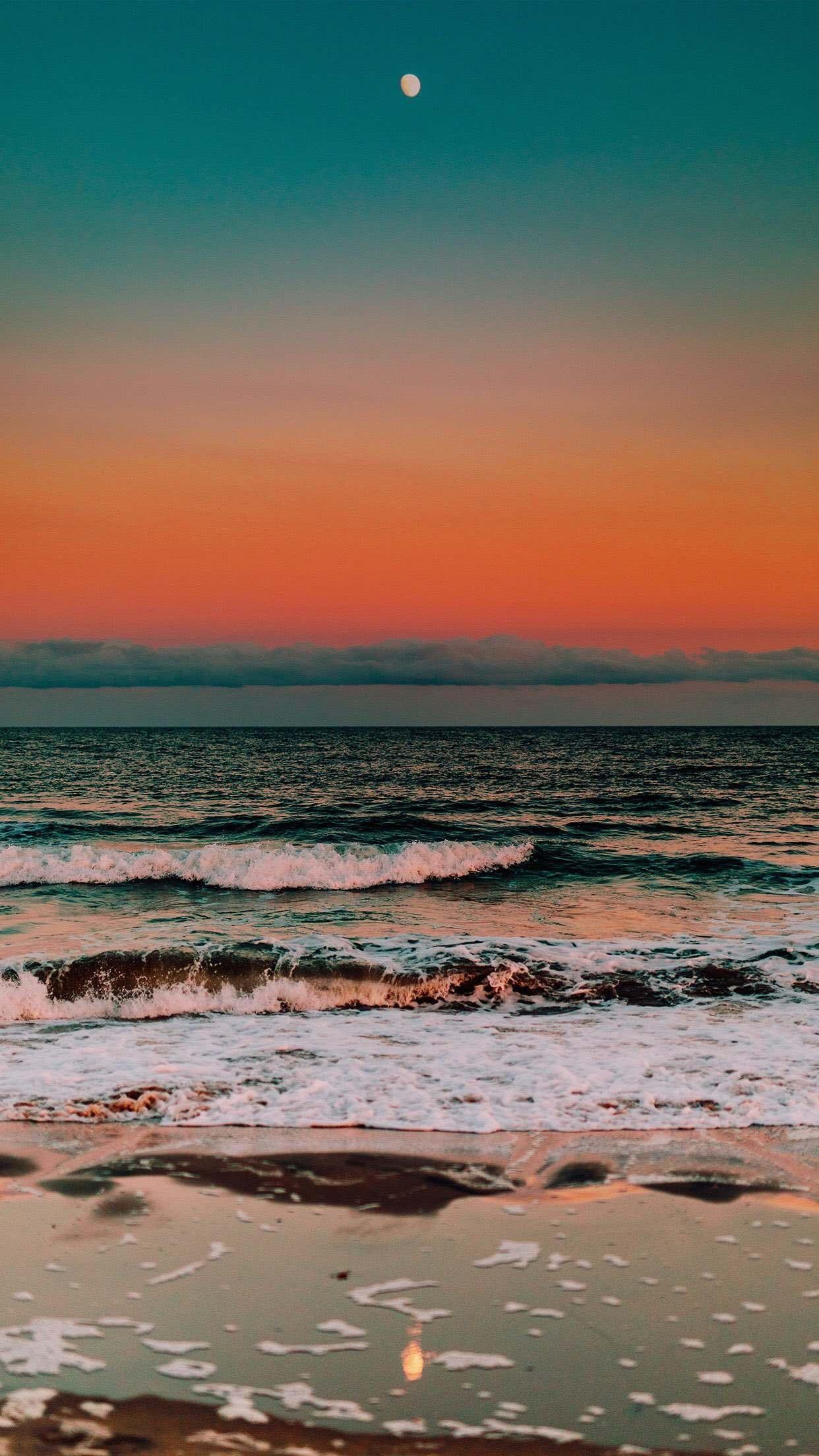 A serene summer beach at sunset with gentle waves lapping the shore, the moon visible in the colorful sky
