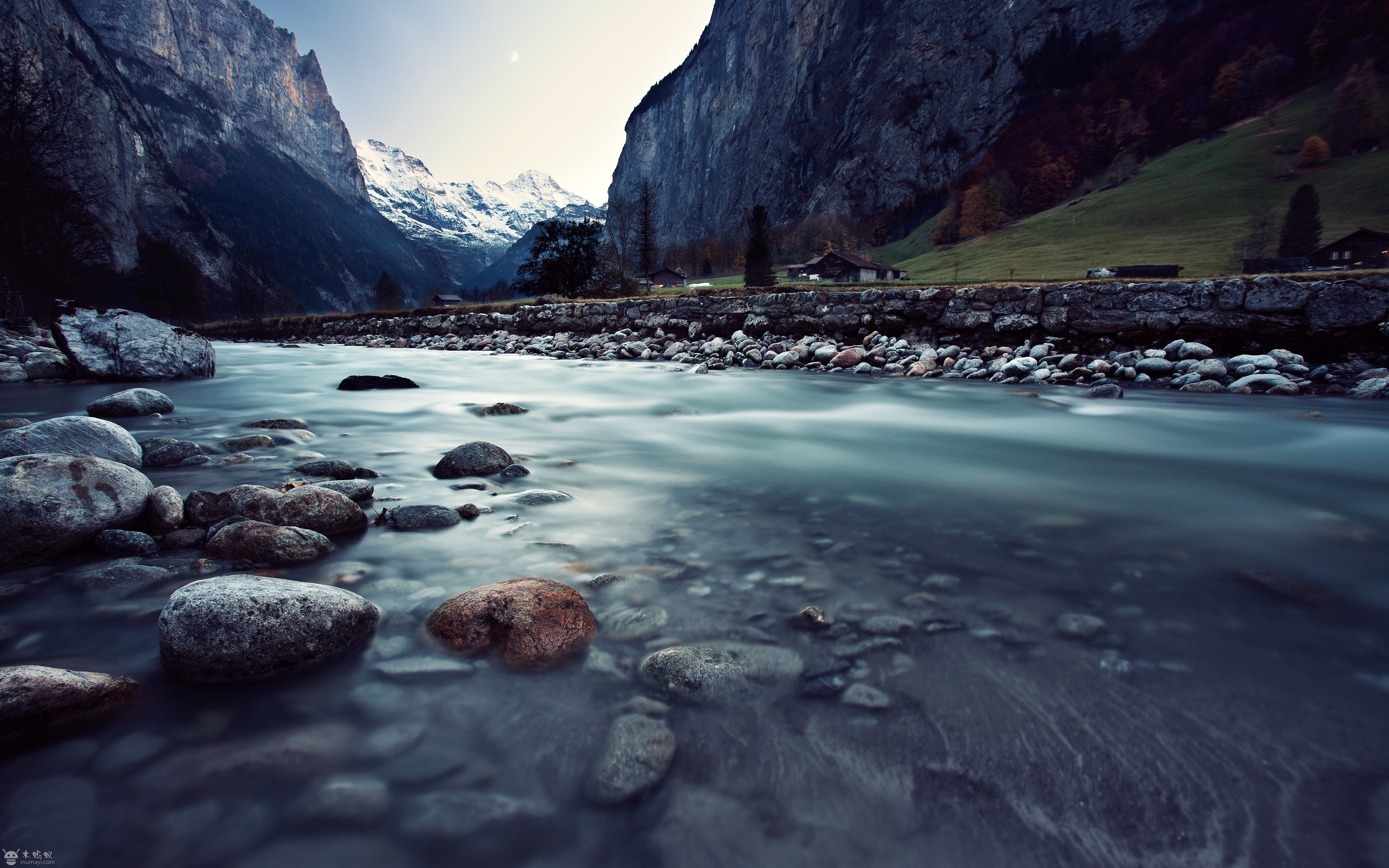Long exposure photo of a pristine river flowing through a valley with snow-capped mountains, rocky riverbed, and lush green hills in the background