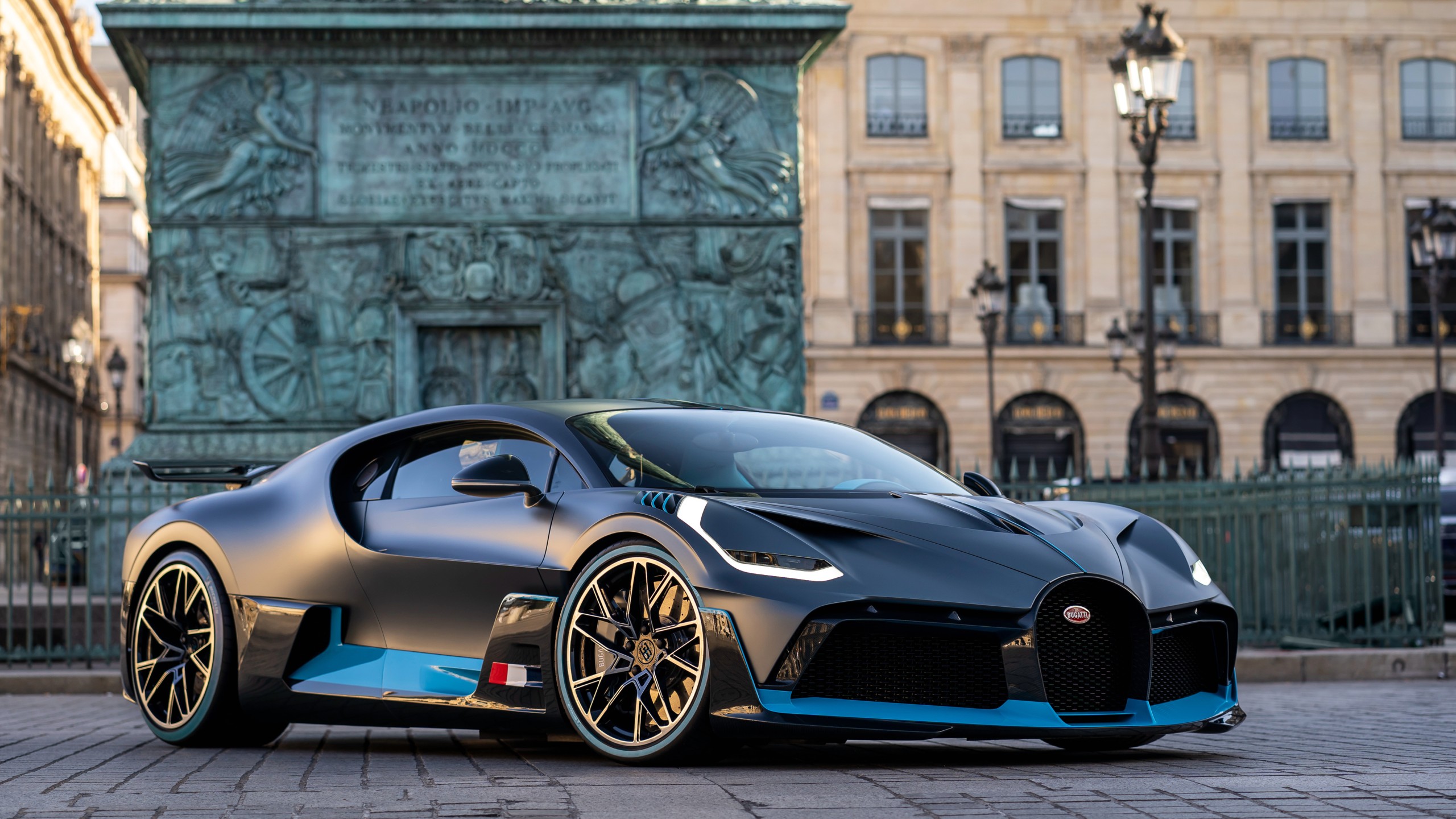 A sleek, dark gray Bugatti Chiron Super Sport, accented with light blue stripes, parked in front of a historical monument in a European city