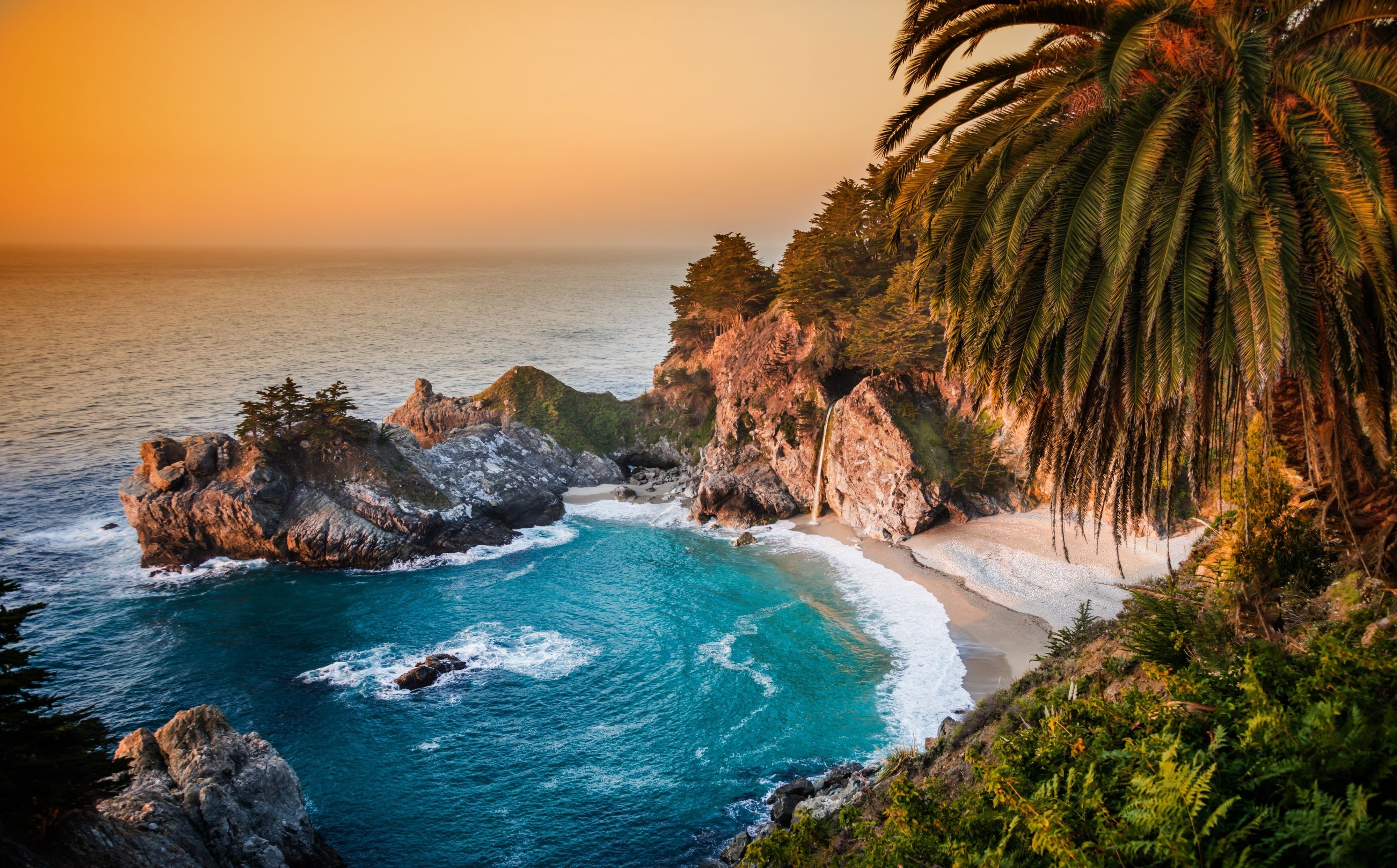 A breathtaking sunset over a secluded cove with a waterfall cascading onto a sandy beach, perfect for a beach computer background