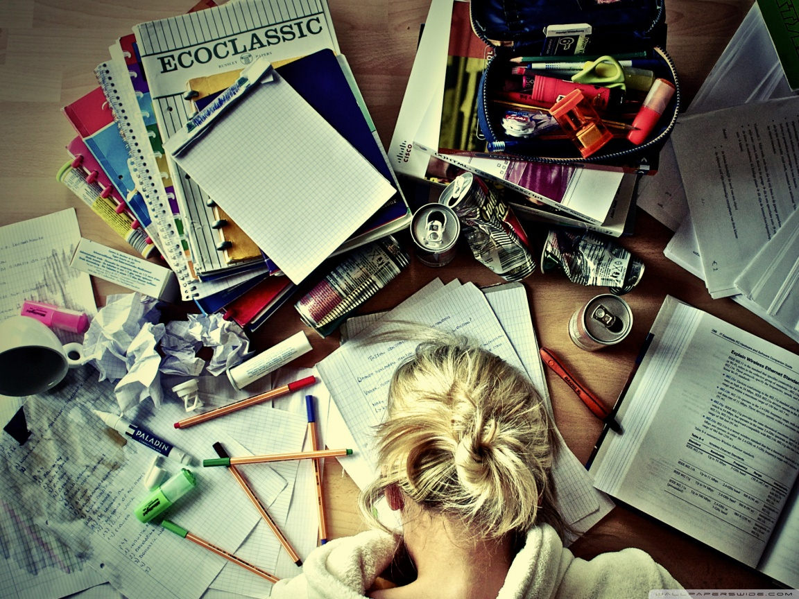 Overhead view of a desk cluttered with study materials, including notebooks, pens, pencils, and crumpled paper, with a person resting their head on the desk, suggesting study fatigue or burnout