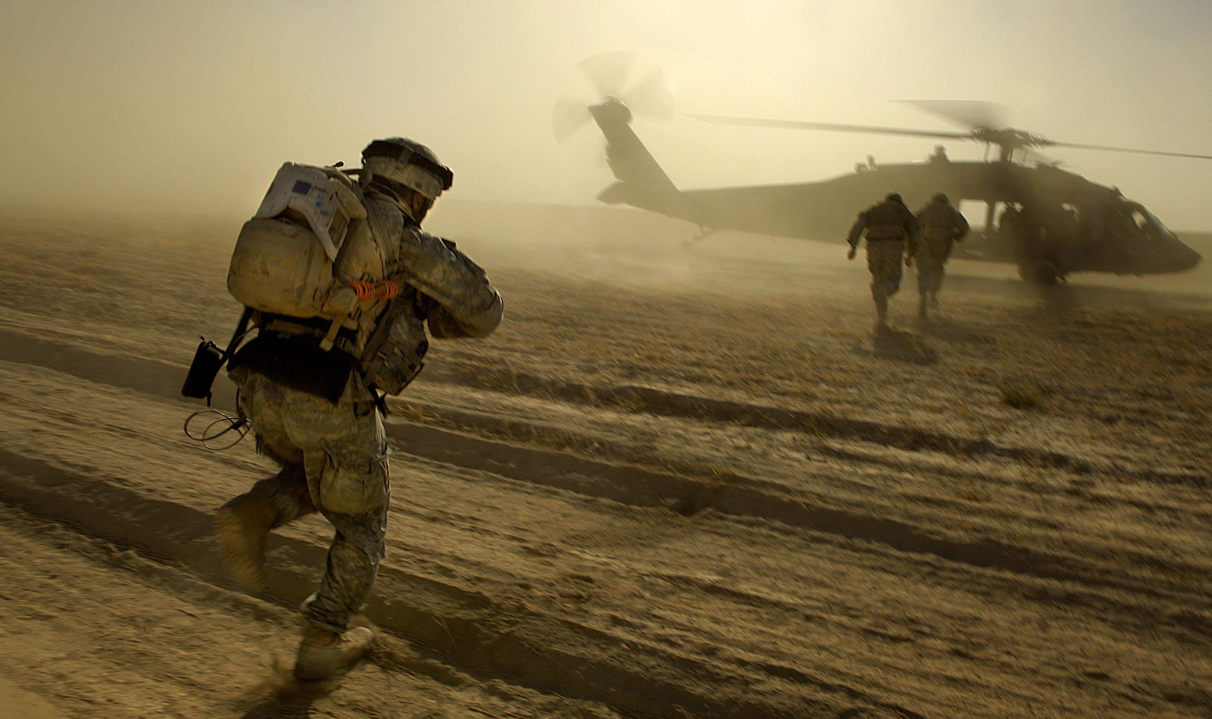 A soldier runs in the desert towards a military helicopter, dust billowing around them
