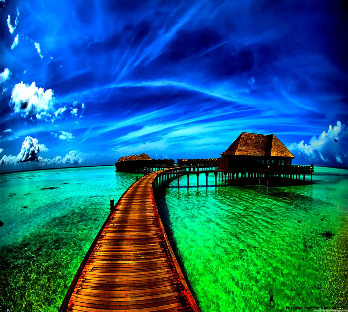 A wooden pier winds its way towards overwater bungalows in a vibrant turquoise ocean, set against a stunning blue sky with wispy clouds. This idyllic tropical paradise scene is perfect for a desktop background