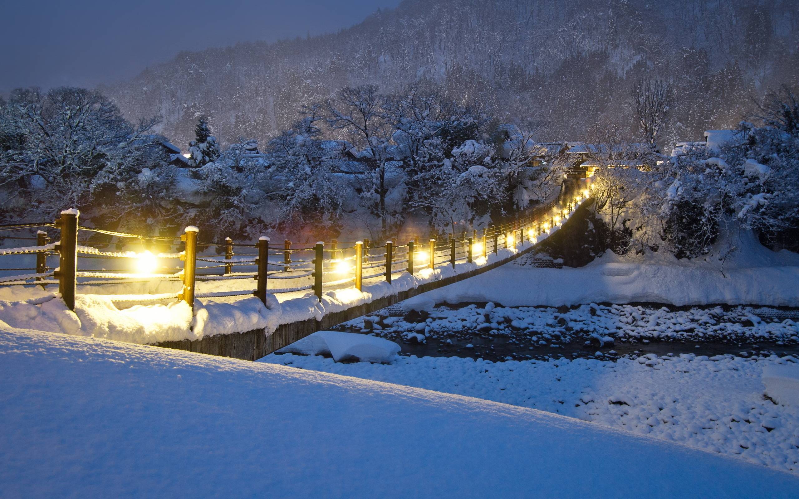 A snow-covered bridge illuminated by warm lights at dusk, leading to a cozy village nestled in a snowy winter landscape
