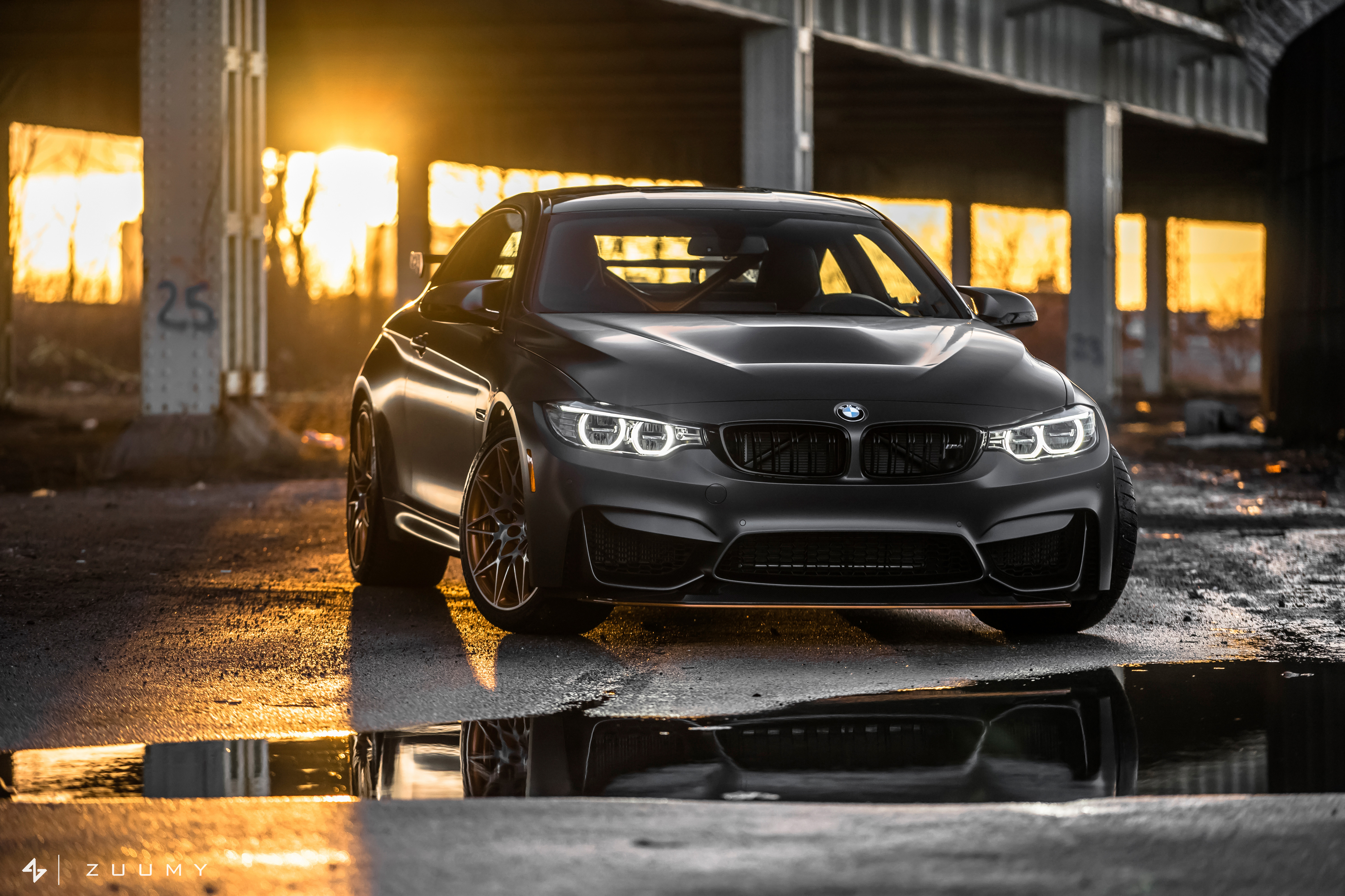 A sleek, black BMW M4 parked under a bridge at sunset, its reflection shimmering in a puddle on the ground