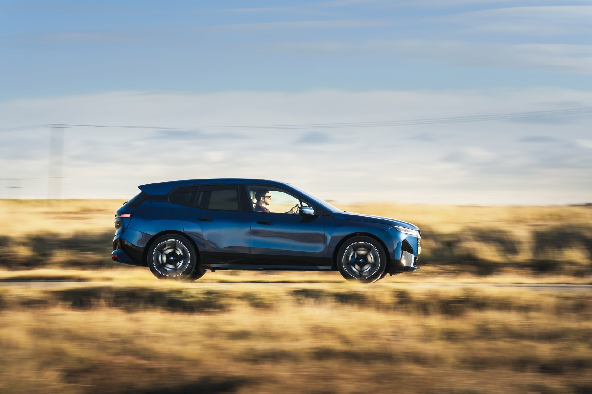 A dynamic side view of the BMW iX electric SUV cruising down a scenic road, showcasing its sleek profile against a backdrop of golden fields and a clear sky