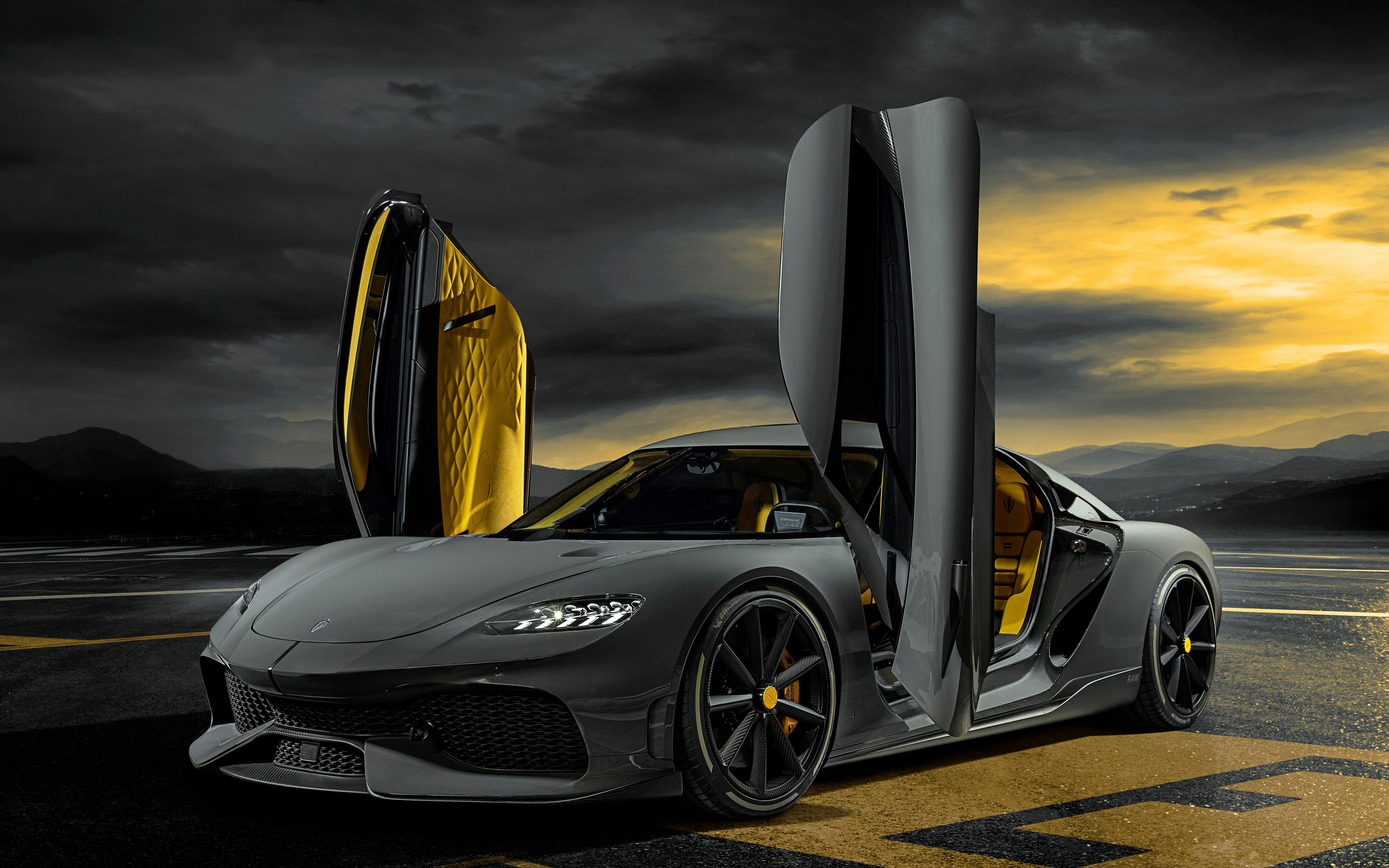 A dark gray Koenigsegg Gemera with gold interior and scissor doors open, parked on a tarmac with a dramatic sky and mountains in the background
