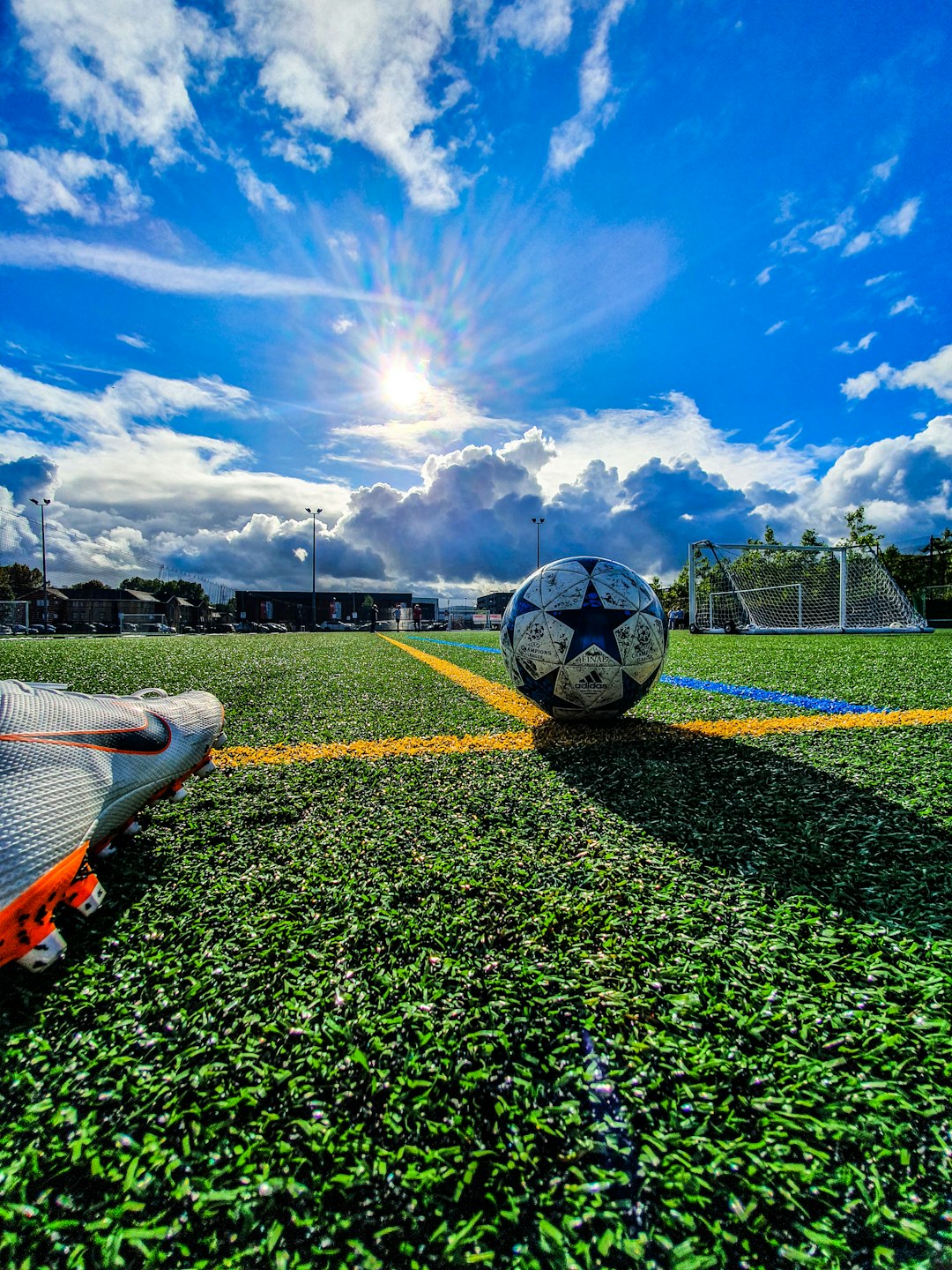 Soccer ball on a vibrant green turf field under a bright blue sky with the sun shining. A soccer cleat is visible in the bottom left corner