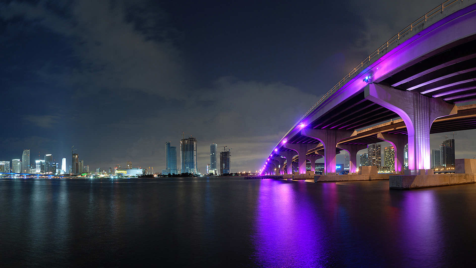 A stunning panoramic view of the Miami skyline at night, with a bridge illuminated in vibrant purple light reflecting on the water