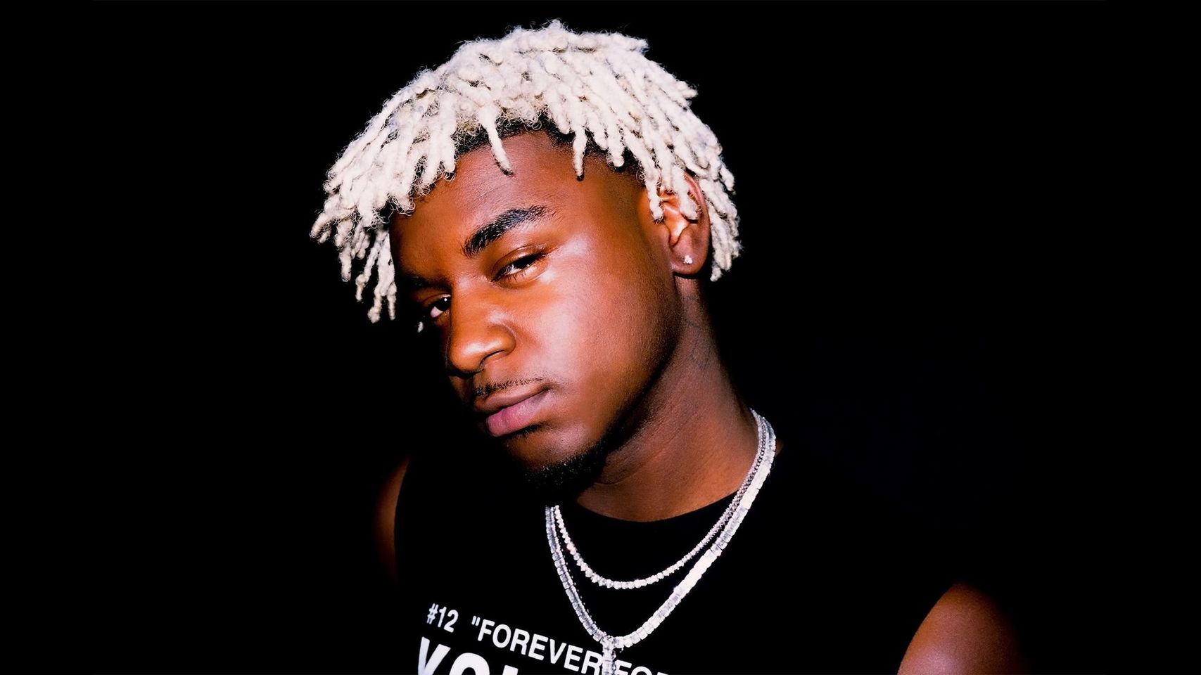 Close-up portrait of rapper Ken Carson against a black background, highlighting his unique hairstyle and jewelry