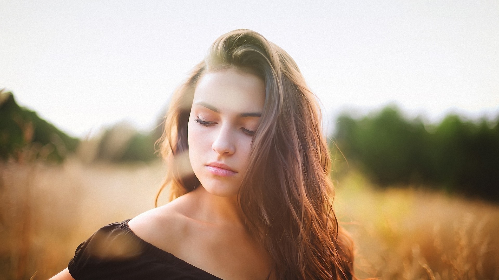 A young woman with long brown hair, wearing an off-the-shoulder black top, stands in a golden field with her eyes closed, conveying a sense of tranquility and peace