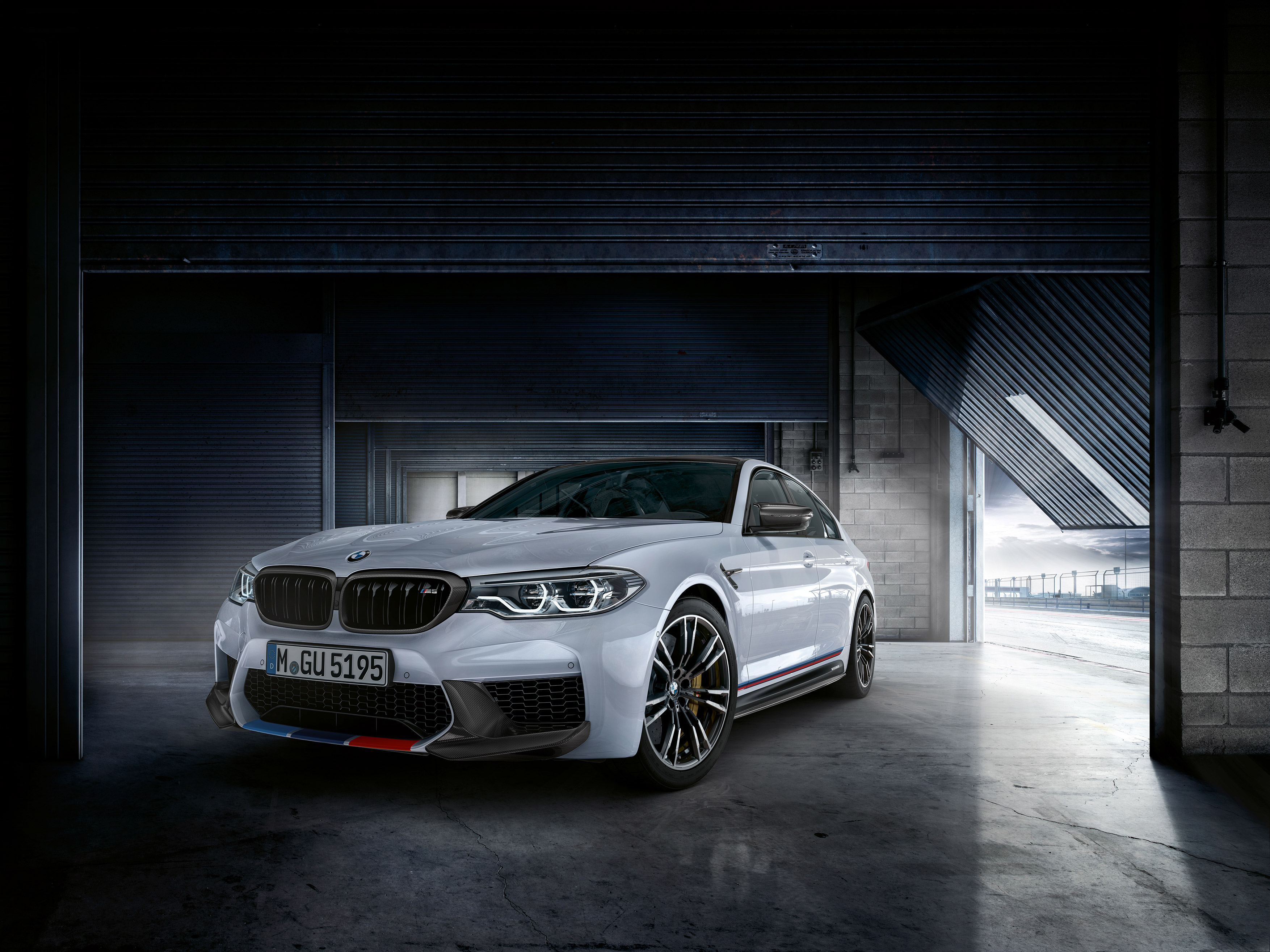 White BMW M5 parked inside a garage, with the garage door partially open, revealing a race track in the background