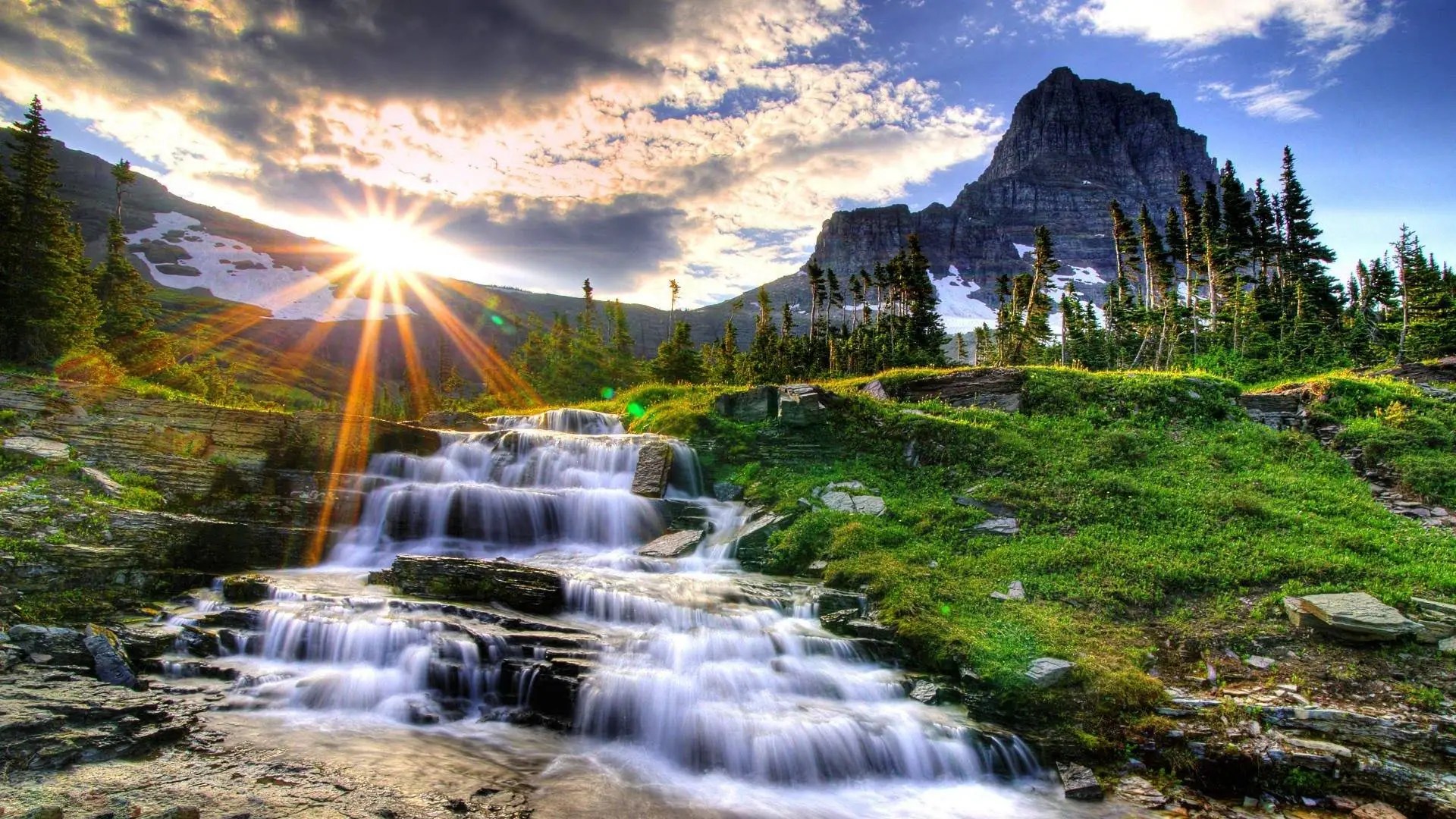 A picturesque waterfall cascades down a rocky slope, illuminated by the warm glow of the setting sun. Lush green vegetation and towering evergreen trees frame the scene, with a majestic mountain peak rising in the background