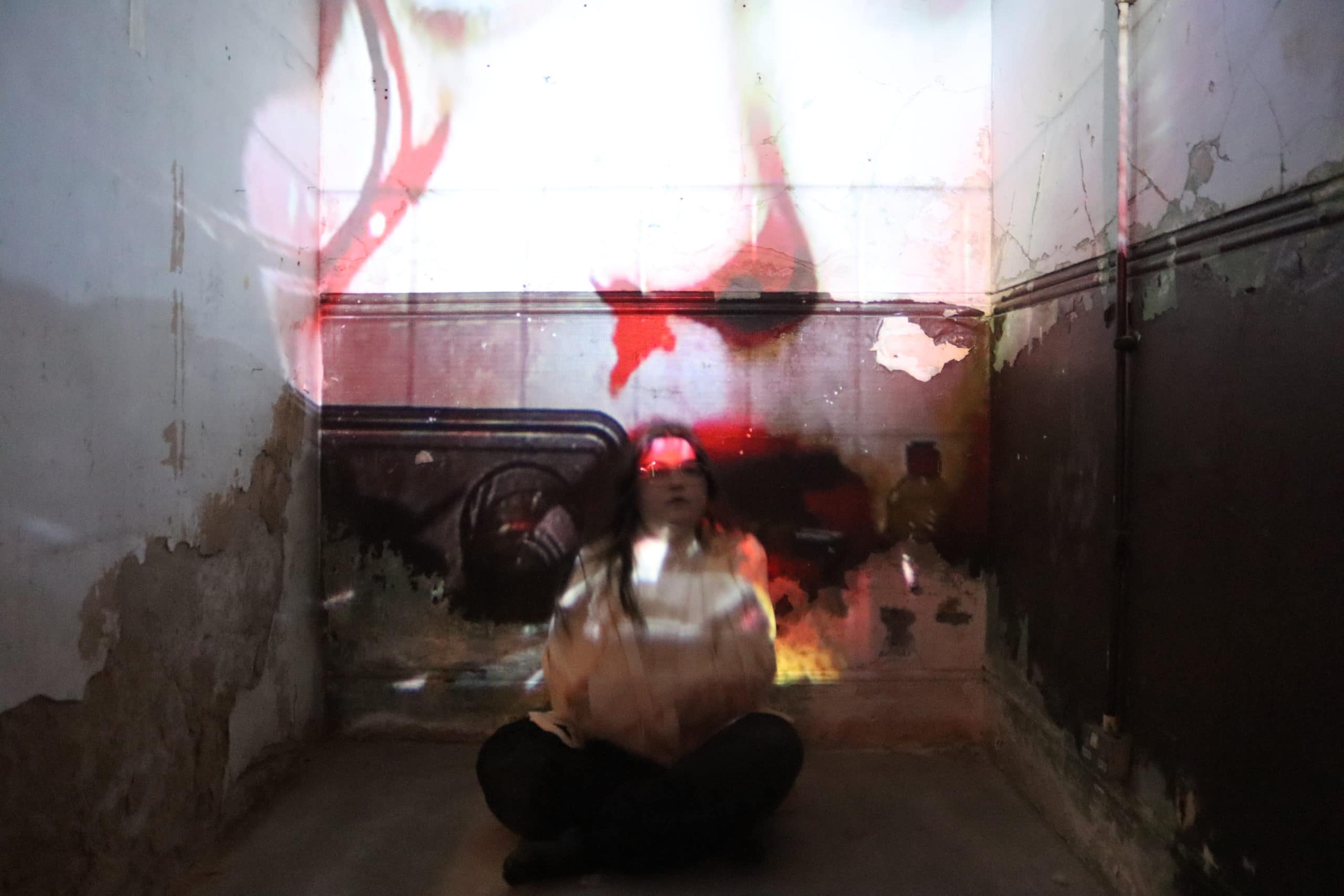 A person sits cross-legged in a dimly lit room, illuminated by a projected image with red and white hues. The walls show signs of age and decay, contrasting with the ethereal nature of the projected light