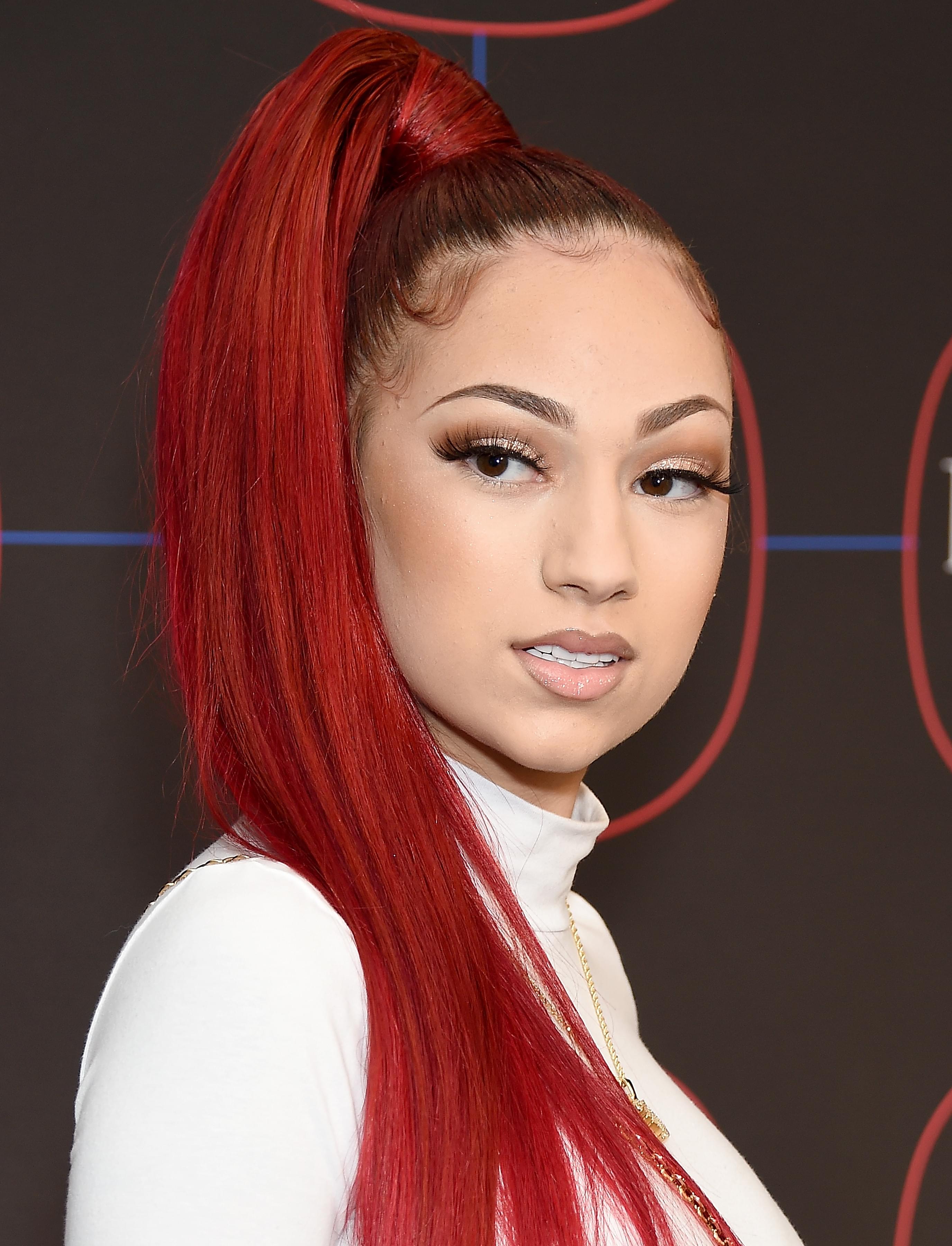 Bhad Bhabie with bright red hair in a high ponytail, wearing a white turtleneck top and gold chain, poses against a black backdrop at an event