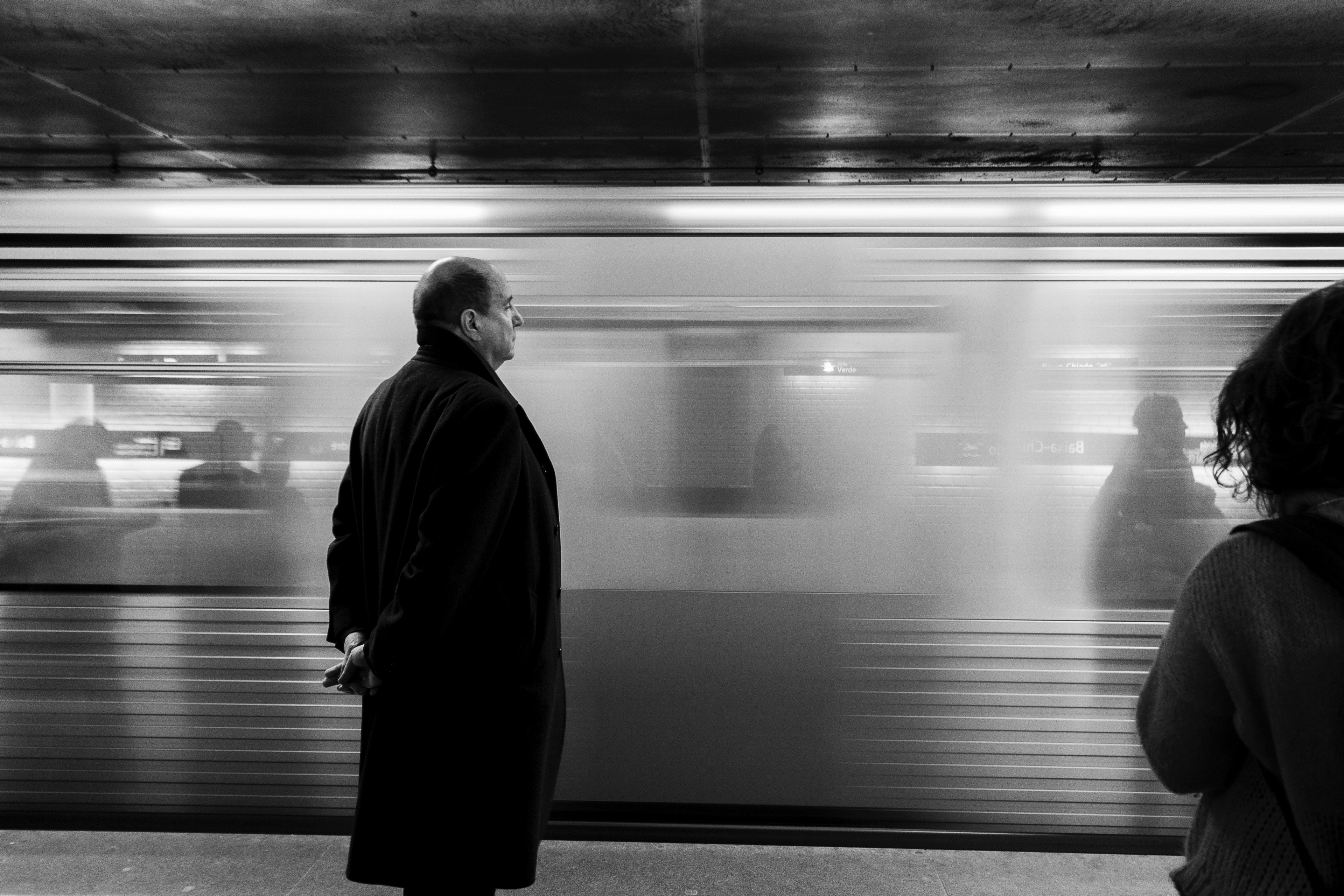 Black And White Shot Of Bald Man Standing In Coat On Subway Platform With Moving Train Wallpaper