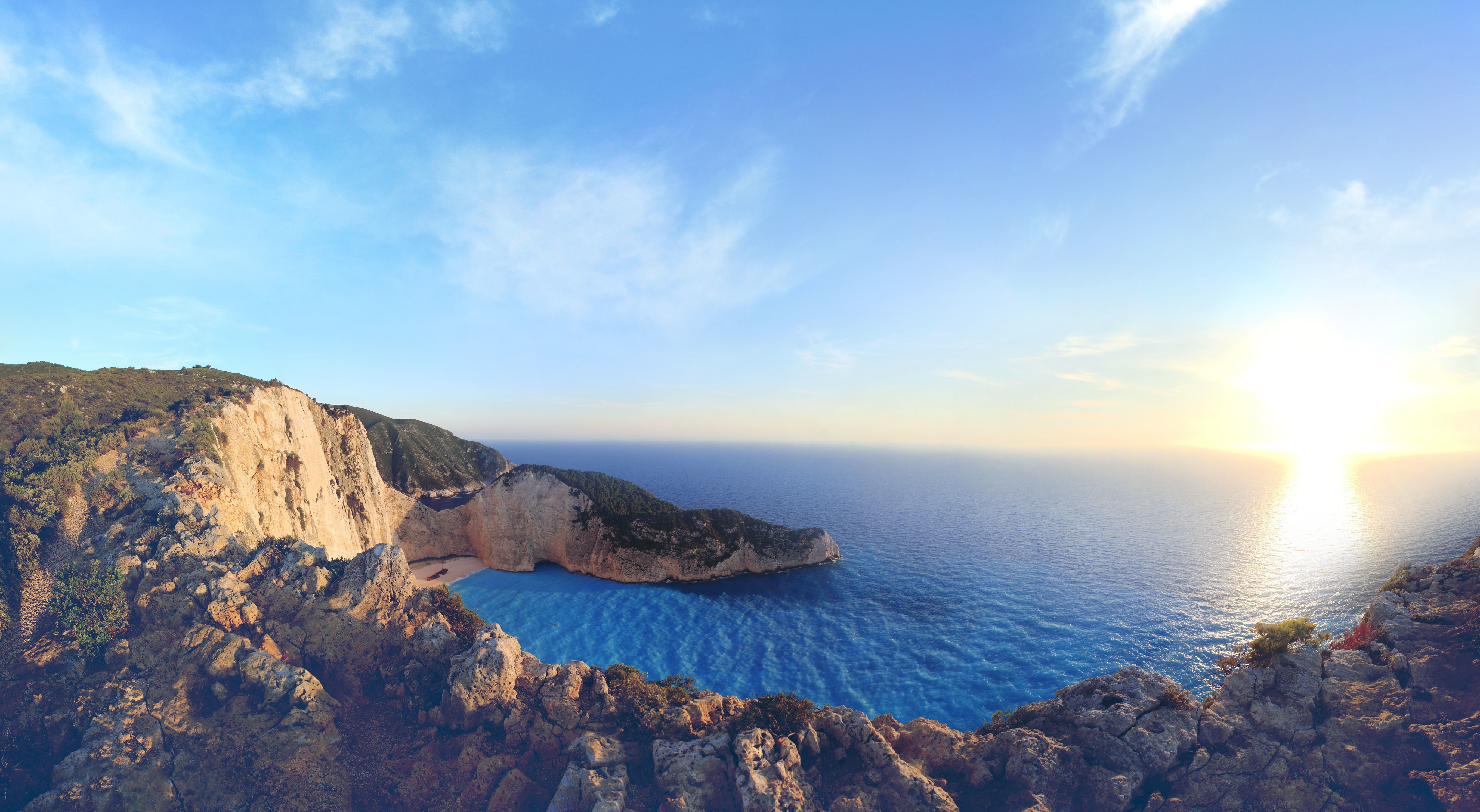 A breathtaking panoramic view of Navagio Beach (Shipwreck Beach) in Zakynthos, Greece, at sunset. The turquoise waters contrast with the steep, rocky cliffs and the setting sun casts a golden glow over the scene