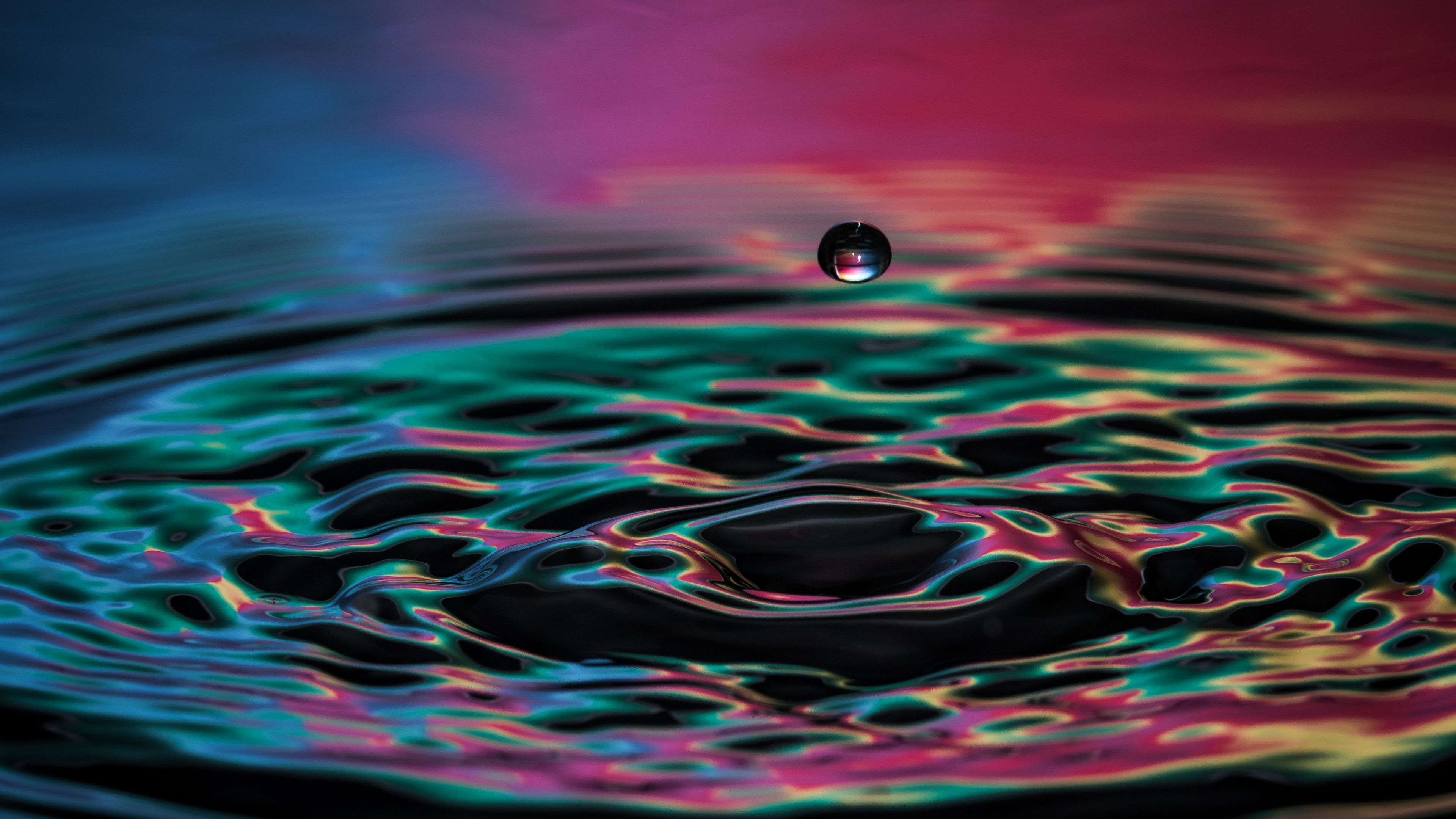 Close-up of a water droplet suspended above a colorful, rippling water surface, creating mesmerizing concentric circles and reflections