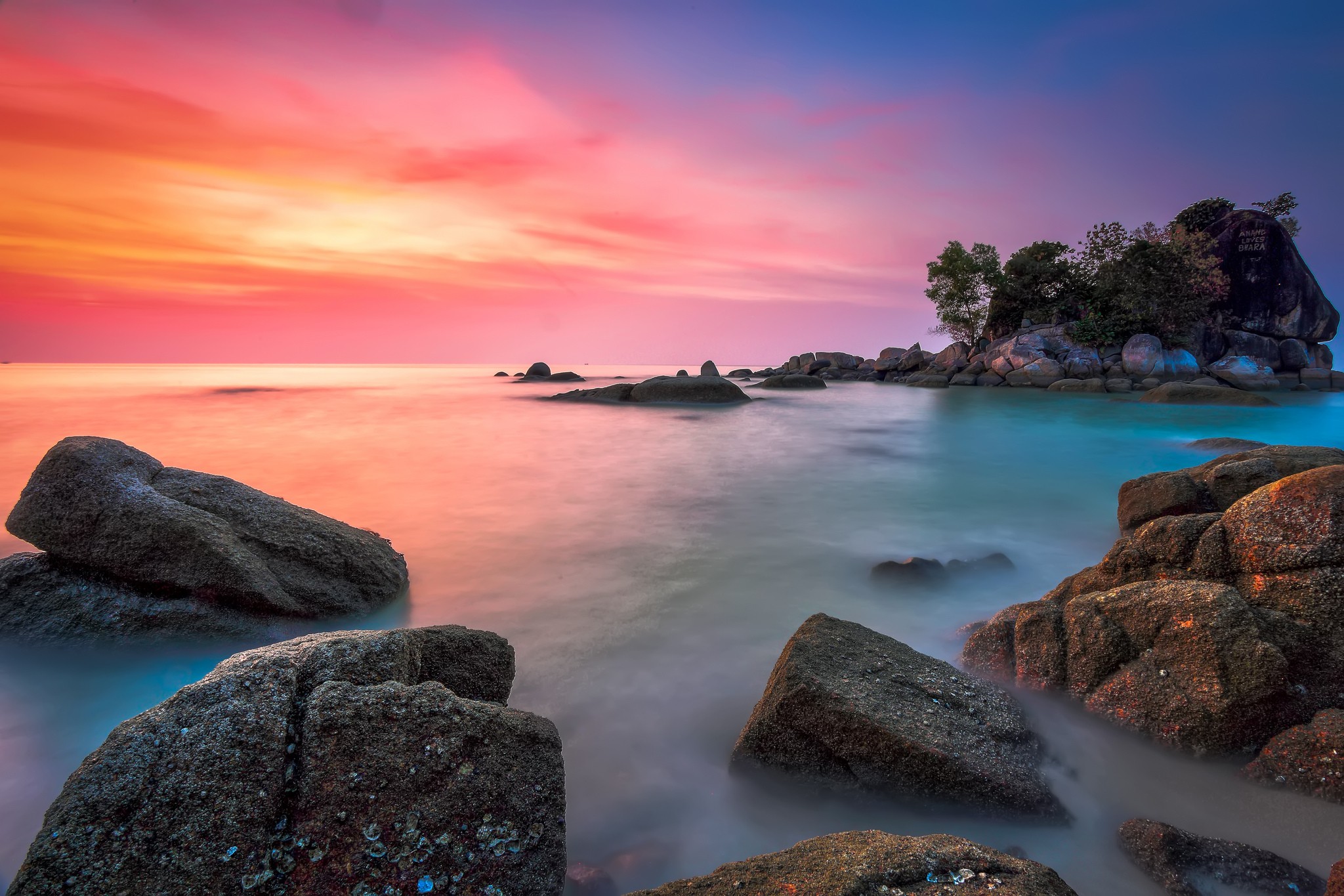 A mesmerizing sunset over a tranquil sea, with smooth rocks in the foreground and a small, vegetated island in the distance. The sky is a blend of vibrant pink, orange, and purple hues, creating a breathtaking coastal scene