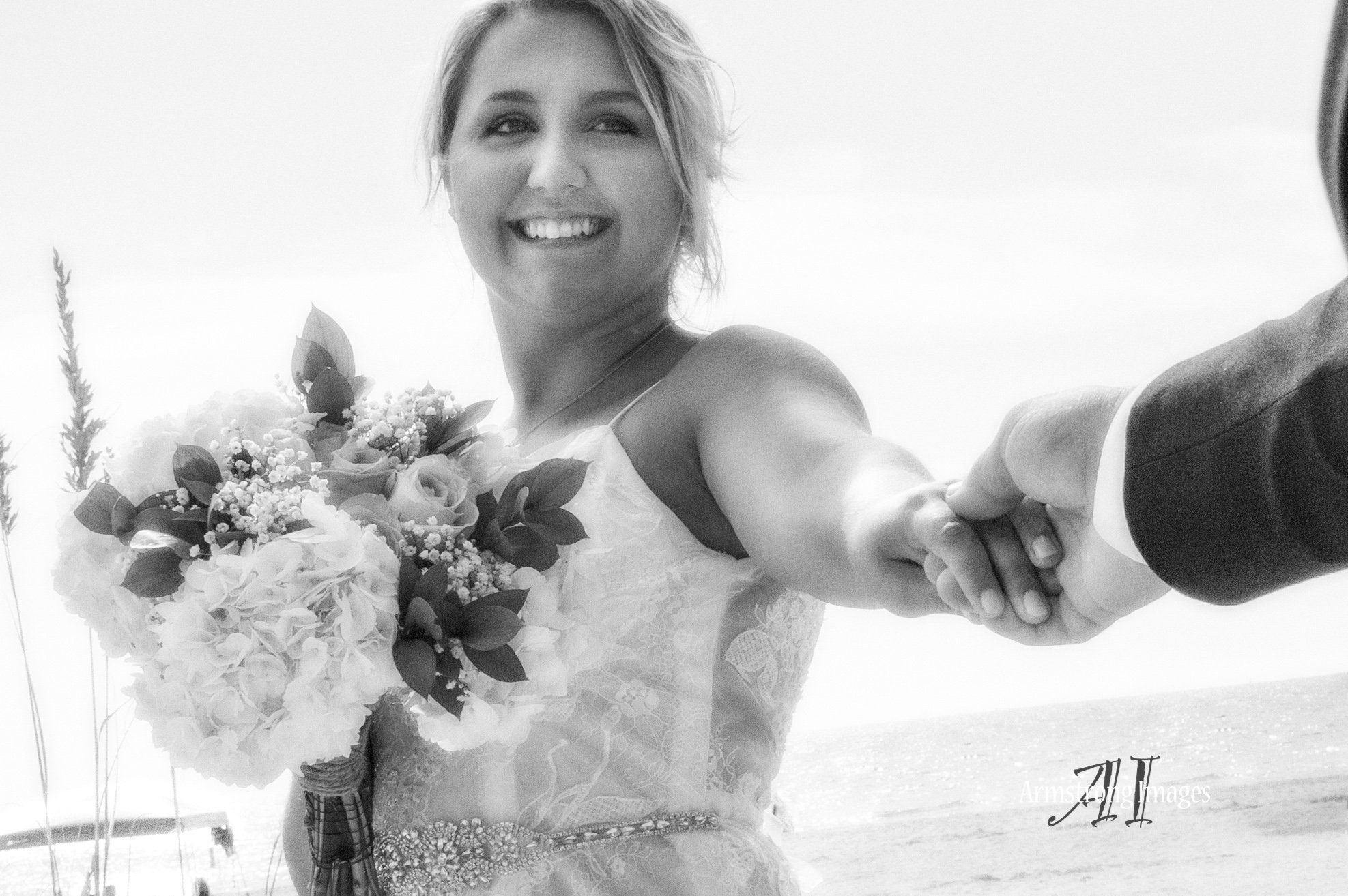Black and white photo of a bride holding a bouquet and her partner's hand, smiling at the camera on a beach