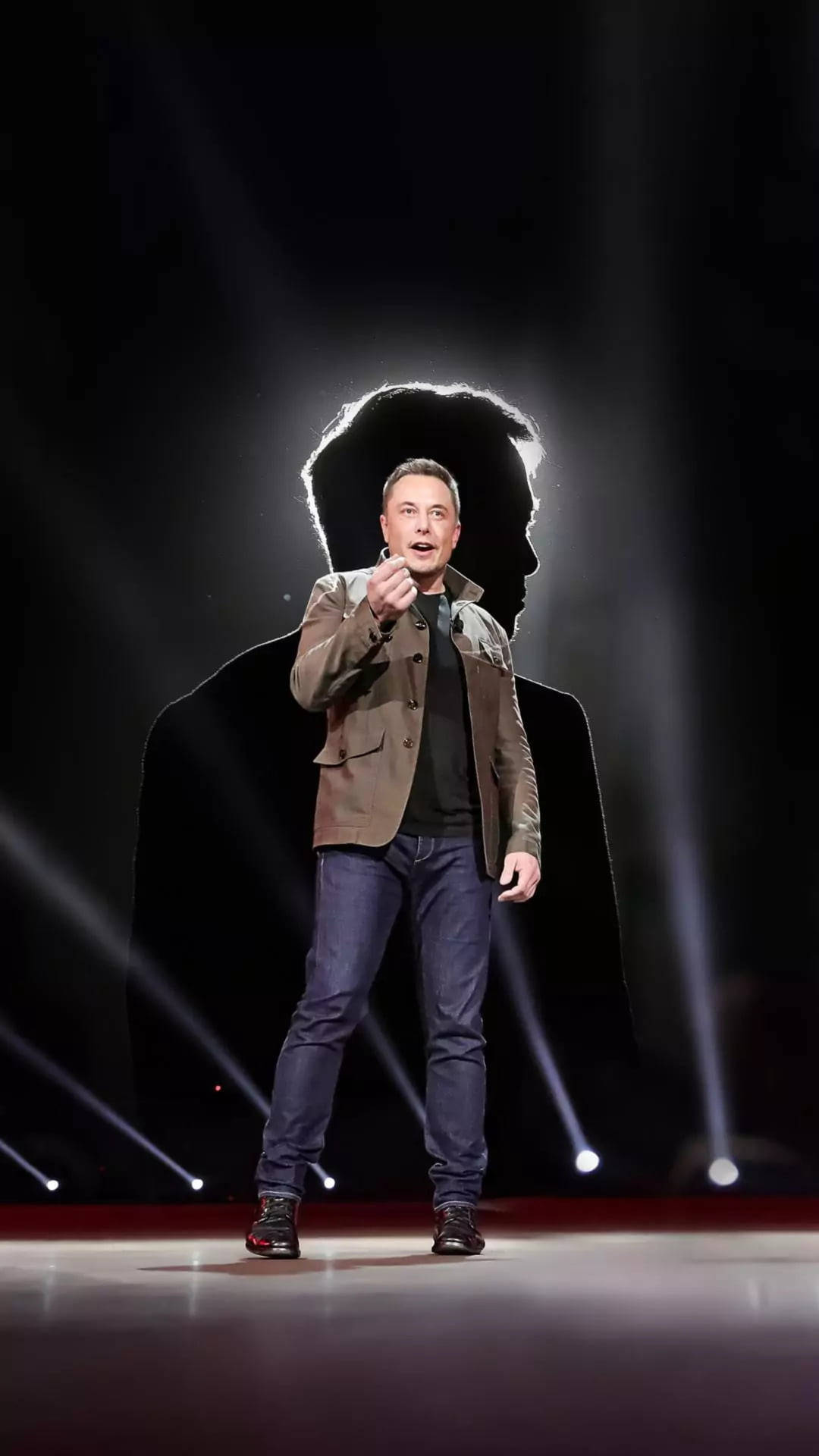 Elon Musk speaking on stage at a Tesla event, with a large shadowy figure of himself behind him